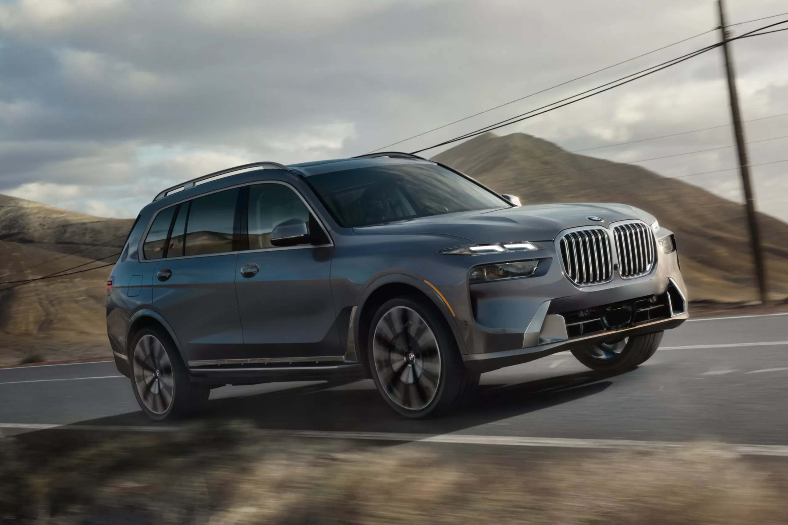 2026 BMW X7 driving on a mountain highway, shown from a front three-quarter angle with bold kidney grille design, large luxury SUV proportions, and performance-focused road presence