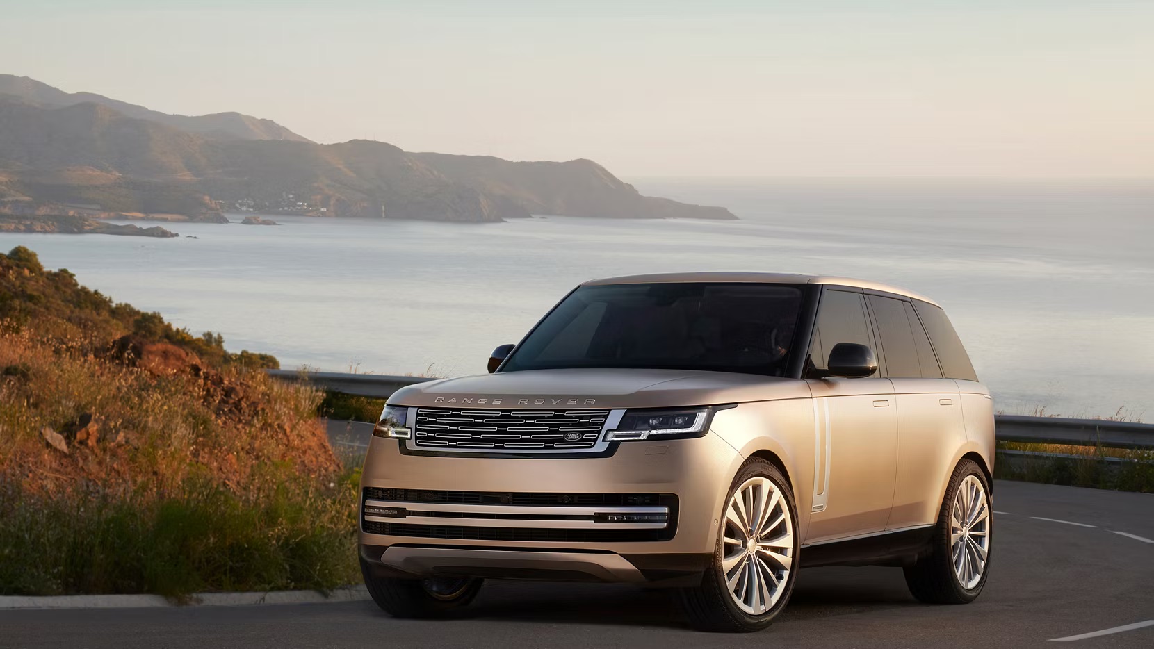 2026 Range Rover driving along a coastal mountain road, shown from a front three-quarter angle with signature luxury SUV styling, panoramic proportions, and upscale road presence
