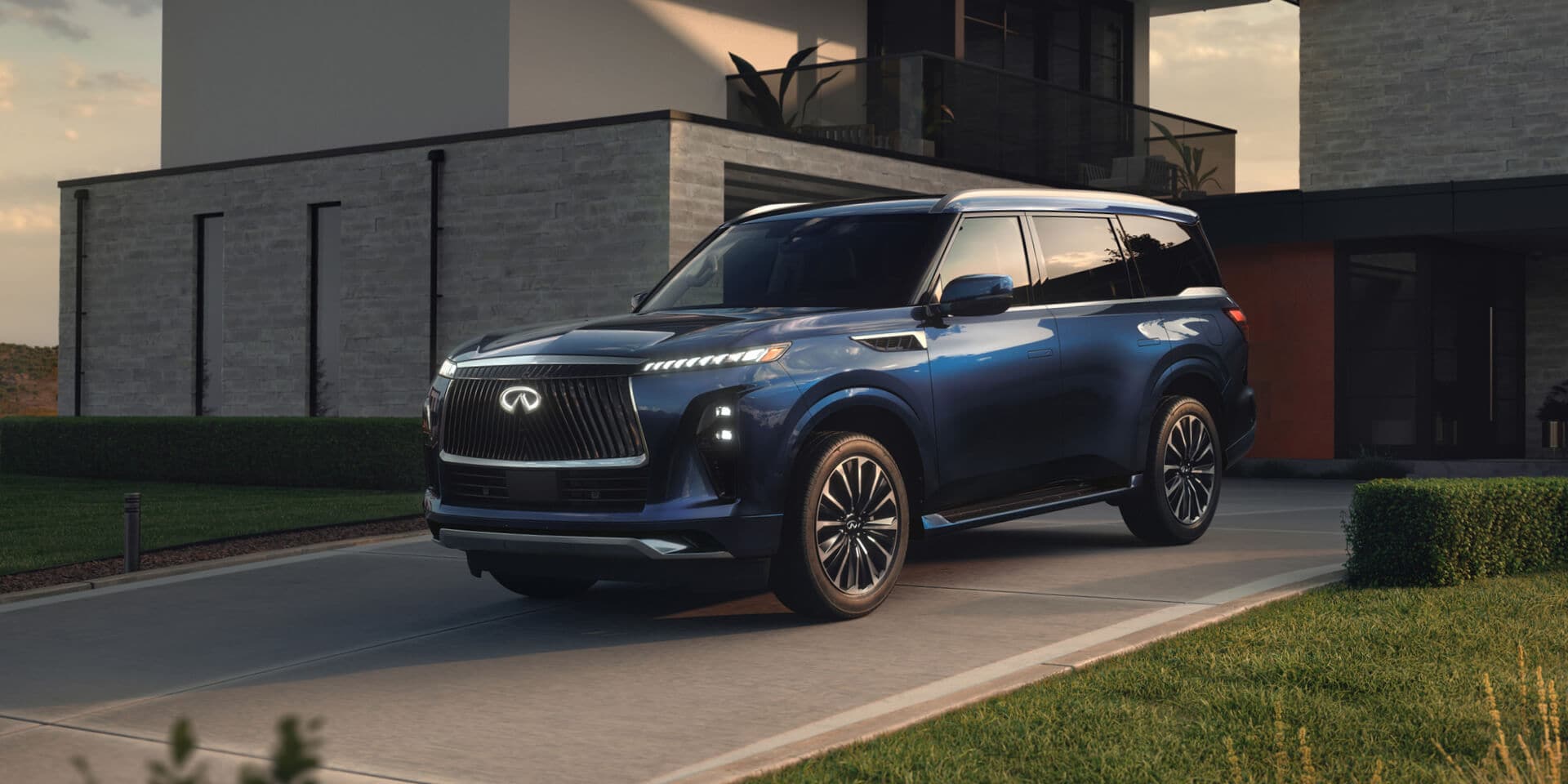 2026 Infiniti QX80 in blue parked in a contemporary residential driveway at dusk, shown from a front three-quarter angle with sculpted body lines, distinctive vertical grille, refined luxury SUV styling, and sophisticated urban presence