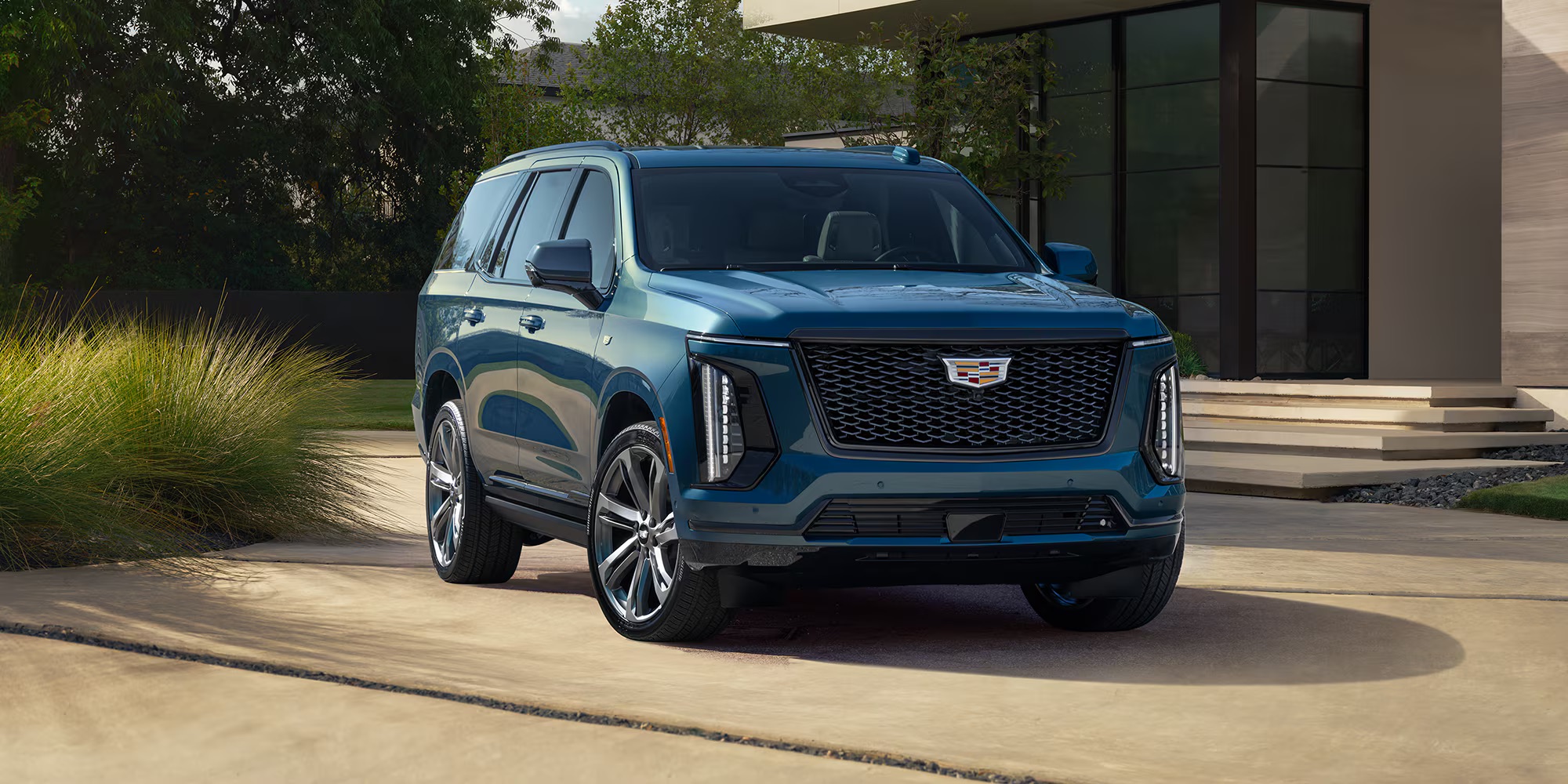 2026 Cadillac Escalade in blue parked outside a modern luxury home, shown from a front three-quarter angle with bold grille design, vertical LED lighting, and full-size premium SUV presence