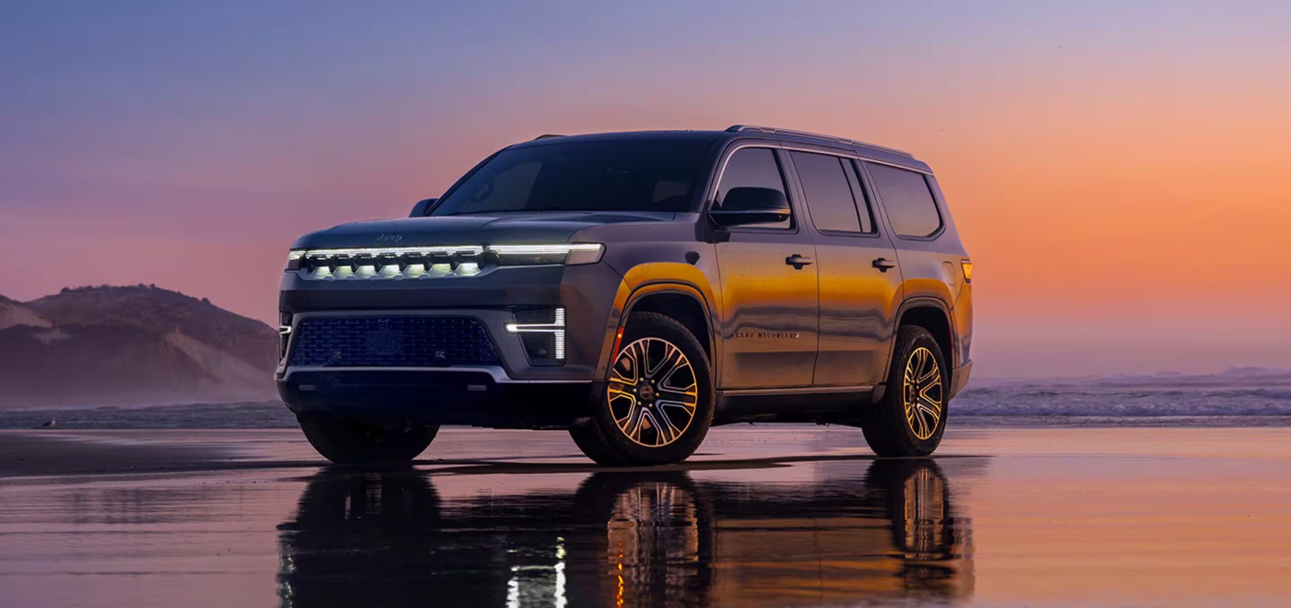 2026 Jeep Grand Wagoneer parked on a beach at sunset, shown from a front three-quarter angle with illuminated grille design, full-size luxury SUV proportions, and upscale American flagship presence