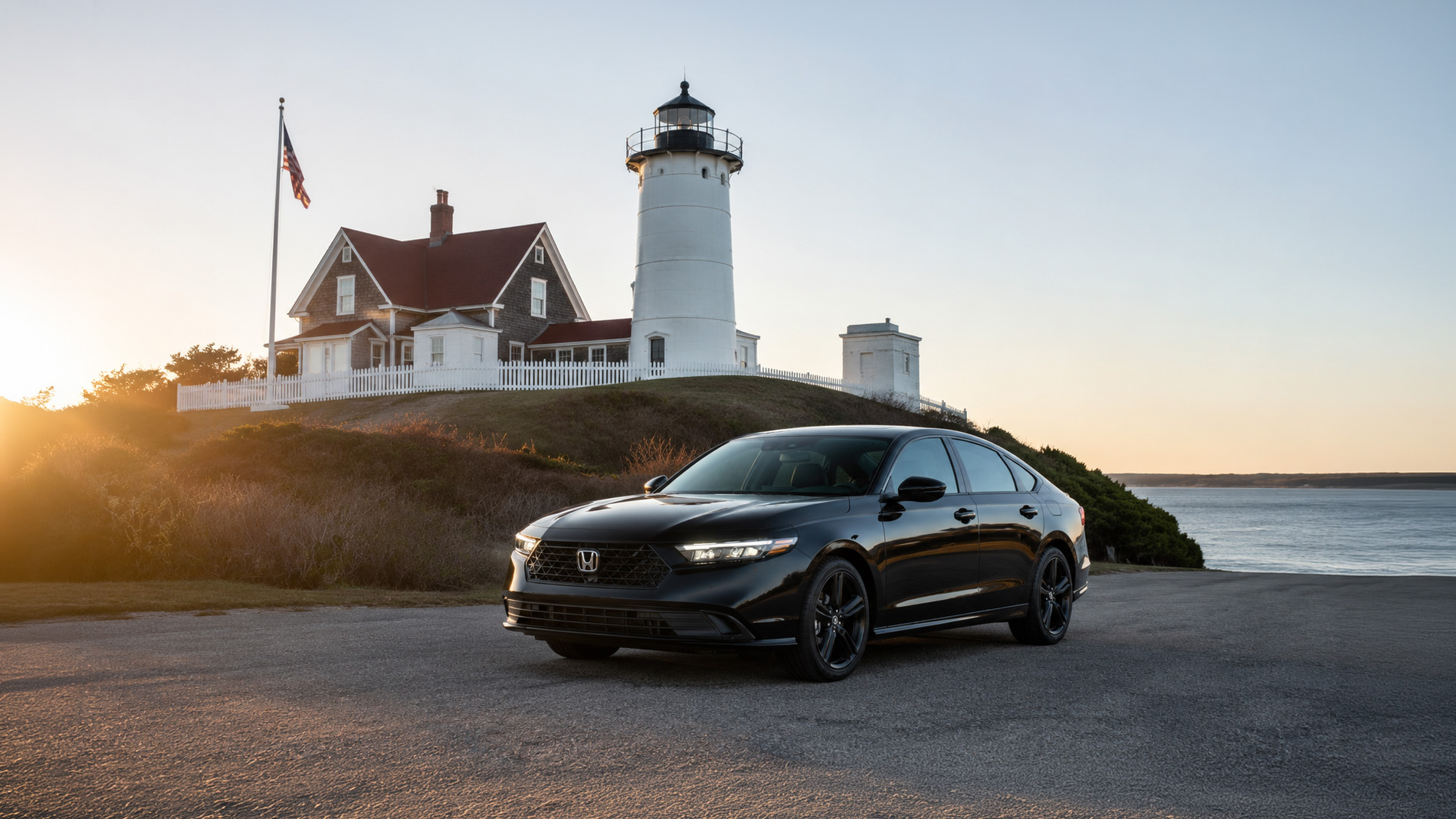 A black 2026 Honda Accord Sport sedan parked near Nobska Lighthouse in Falmouth, Massachusetts at sunset, with the historic white lighthouse, red‑roofed keeper’s house, and American flag in the background overlooking the Atlantic Ocean, showcasing Accord’s sleek design and New England heritage connection.