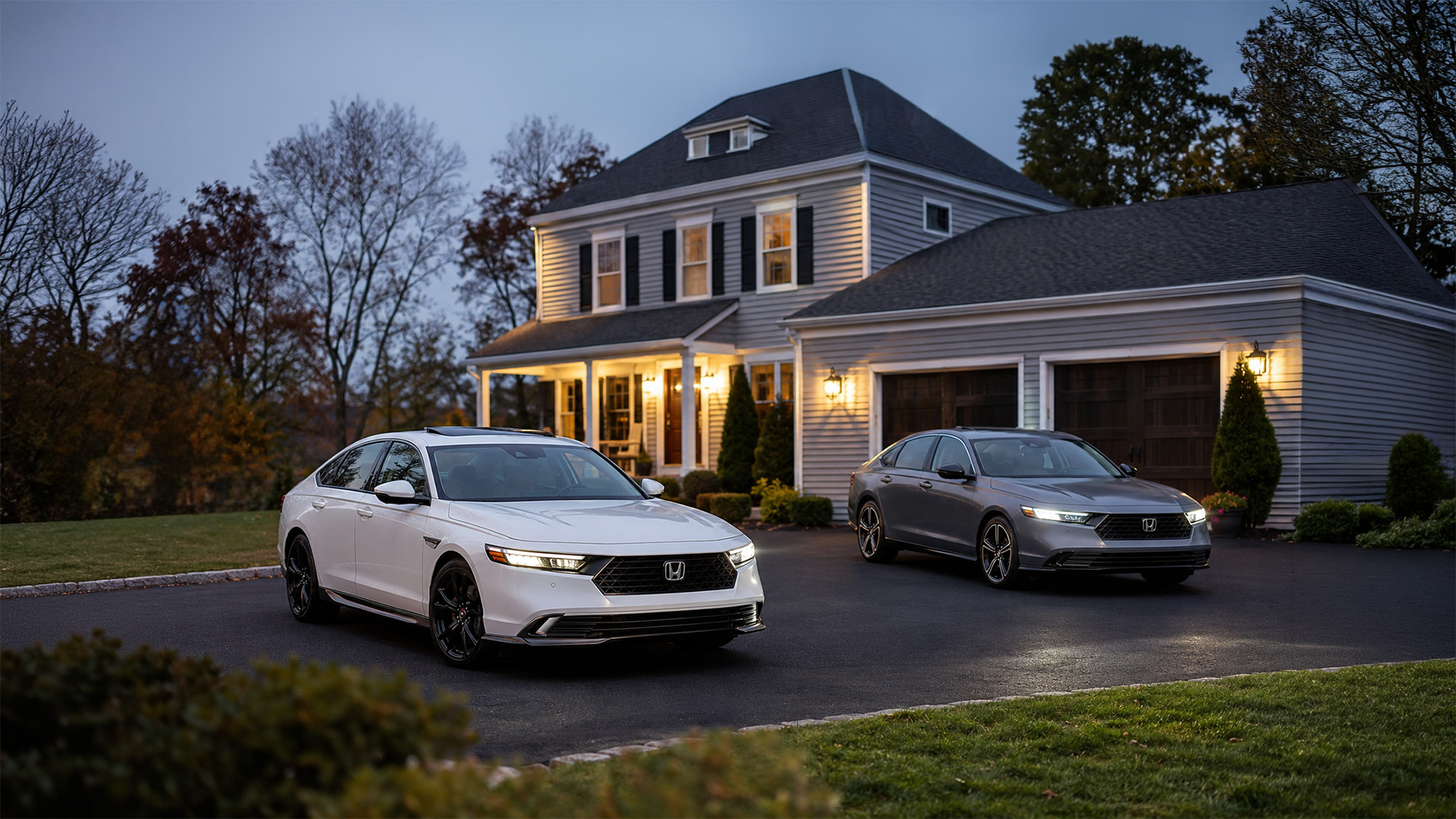 A white and gray 2026 Honda Accord parked side by side in a residential driveway at dusk, with warm porch lights illuminating a two‑story home and autumn trees in the background, showcasing Accord Sport and Touring trims available at Silko Honda in Raynham, MA.