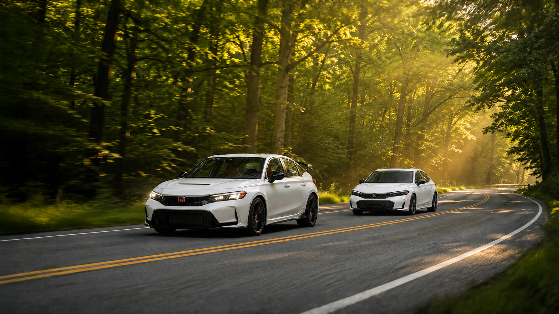 Two white 2026 Honda Civic models drive along a winding forest road at sunrise, with sunlight streaming through the trees and illuminating the roadway, showcasing the sporty Type R and Si trim levels available at Silko Honda in Raynham, MA.