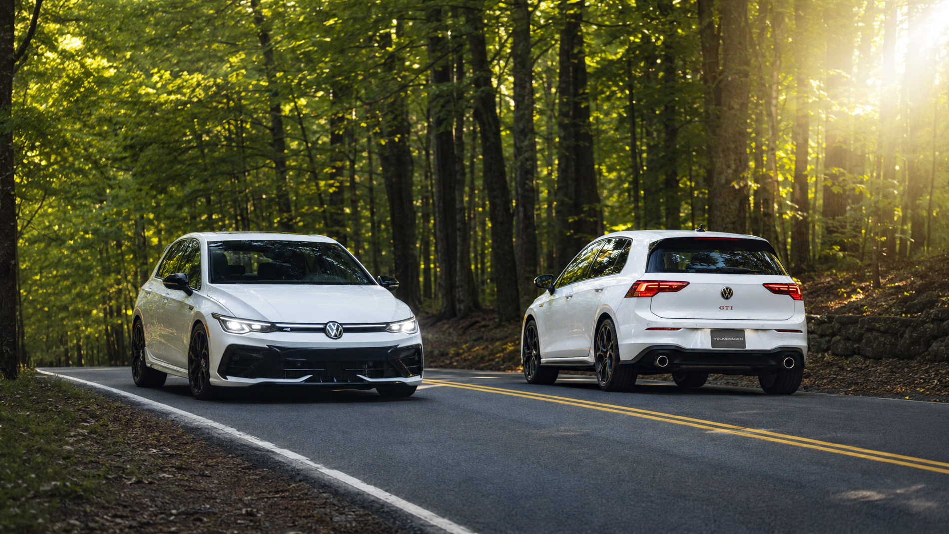 Two white 2026 Volkswagen Golf hatchbacks drive along a winding forest road surrounded by tall trees and soft morning light, illustrating the performance and refined styling of the GTI and R trims.