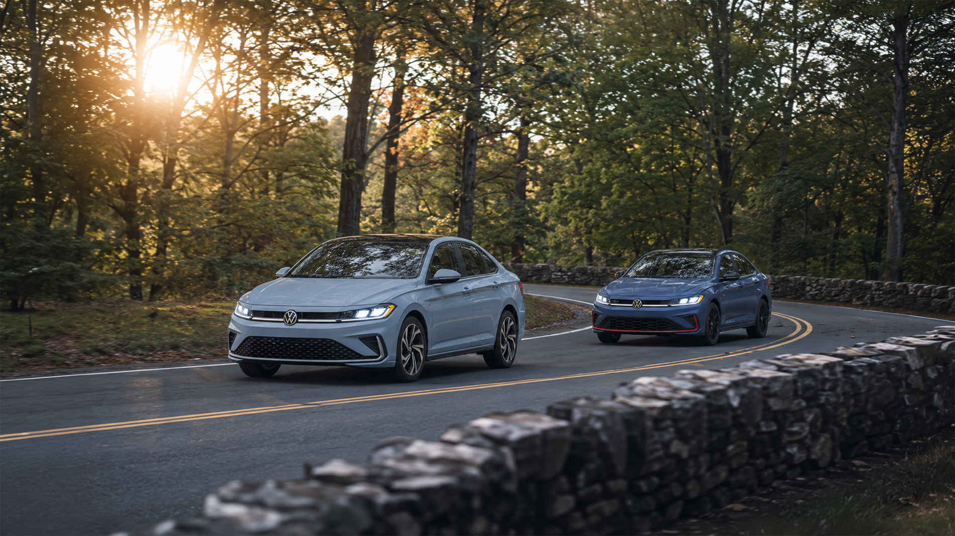Two 2026 Volkswagen Jetta sedans in gray and blue driving along a winding forest road bordered by stone walls and autumn trees, illustrating the contrast between standard and GLI performance trims.
