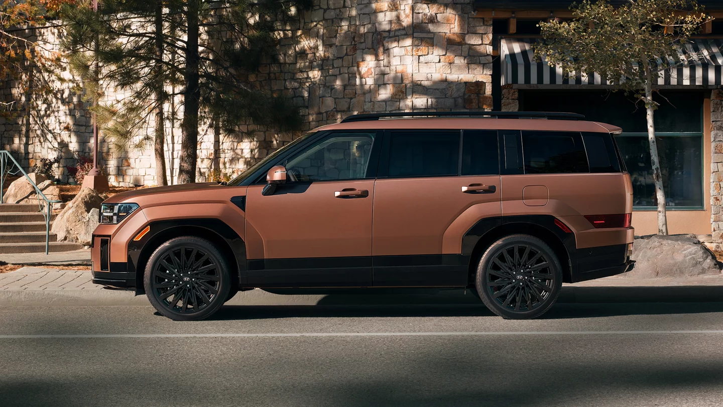 2026 Hyundai Santa Fe parked on a city street with a bronze exterior, boxy SUV styling, black alloy wheels, full-size three-row proportions, and a modern upscale design shown in profile beside a stone building backdrop.