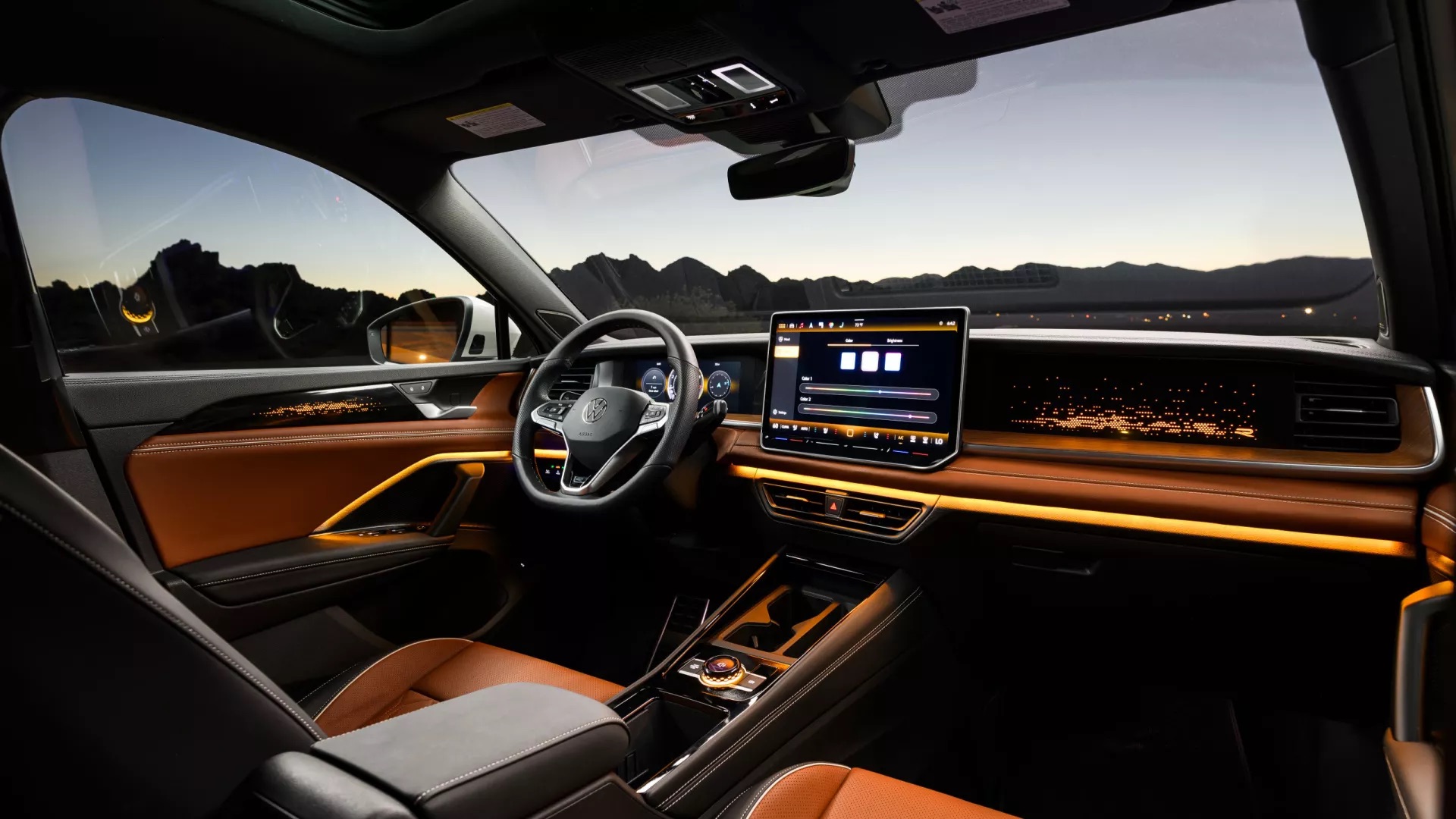 2026 Volkswagen Tiguan interior featuring brown leather seating, illuminated ambient lighting across the dashboard and door panels, a large touchscreen infotainment display, digital driver cluster, and panoramic mountain views visible through the windshield at dusk.
