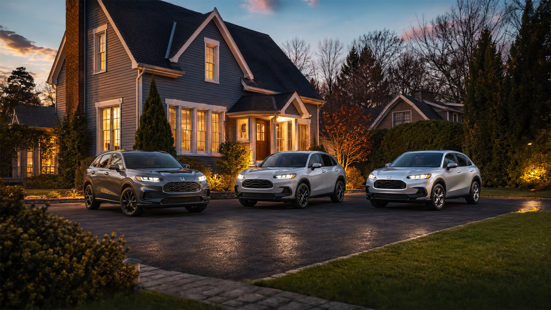 Three 2027 Honda HR-V SUVs in gray and white parked in a residential driveway at dusk, with warm house lights and trees in the background, highlighting the SUV’s refined styling and trim options for suburban drivers.