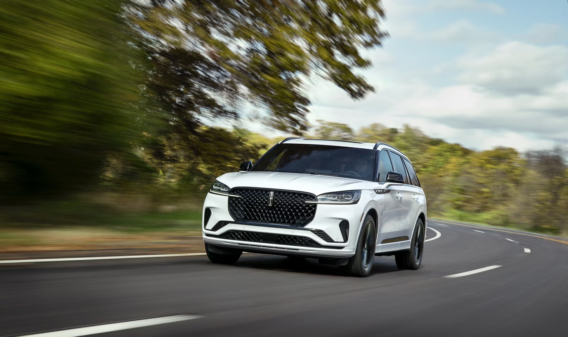 2026 Lincoln Aviator driving on a winding road with a white exterior, updated Lincoln grille design, black roof accents, and slim LED lighting signature shown in motion against a wooded backdrop.