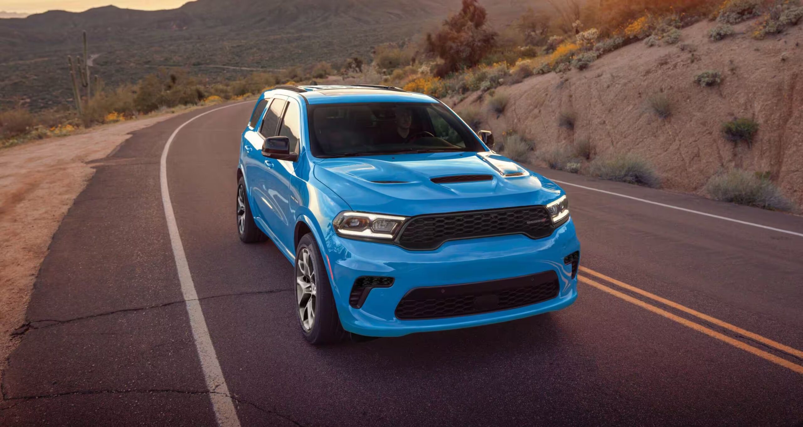 2026 Dodge Durango driving on a winding desert road with a bright blue exterior, hood vents, black performance grille, LED headlights, and muscular three-row SUV styling shown at sunset in a mountainous landscape.