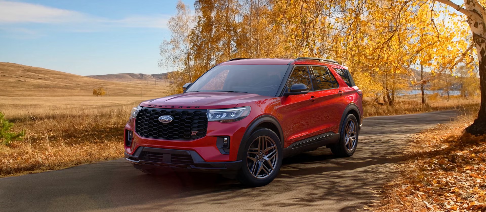 2026 Ford Explorer ST parked on a tree-lined road during autumn with a red exterior, black mesh grille, sporty ST styling, large alloy wheels, and golden fall foliage surrounding the midsize three-row SUV.