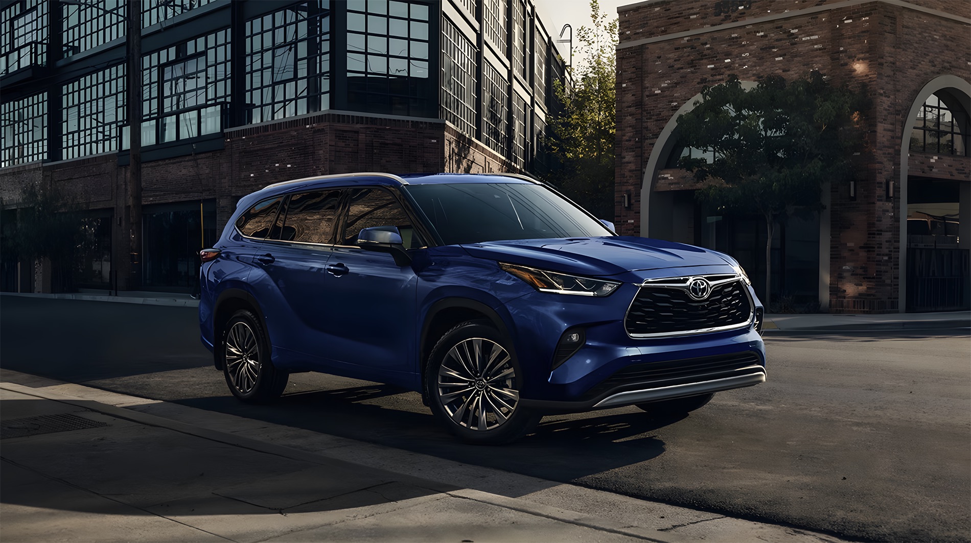 2026 Toyota Highlander parked on a city street at dusk with a blue exterior, large black grille, slim LED headlights, and upscale three-row SUV styling set against modern urban architecture.