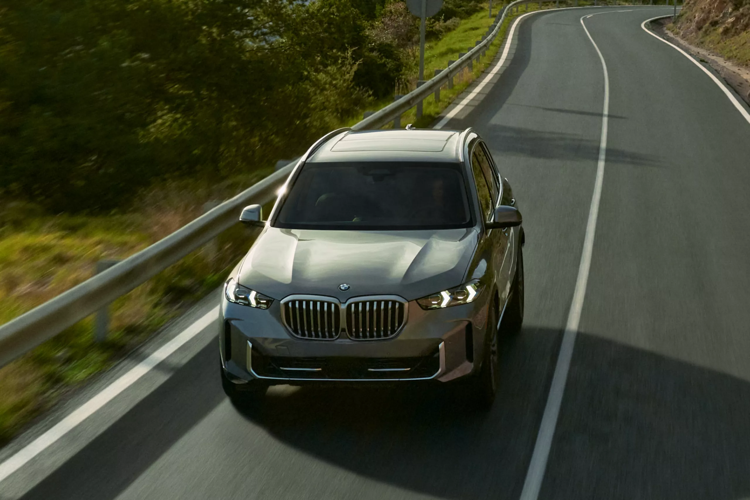 2026 BMW X5 driving on a winding two-lane road with a gray exterior, illuminated LED headlights, signature BMW kidney grille, and midsize luxury SUV styling shown from an elevated front angle.