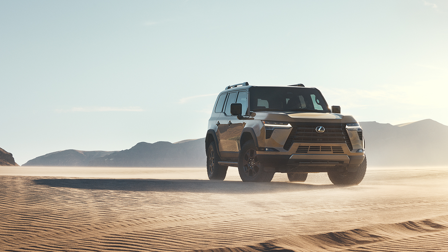 2026 Lexus GX parked on desert sand dunes with a bronze exterior, upright off-road SUV styling, black spindle grille, rugged all-terrain tires, and mountain scenery in the background at sunset.
