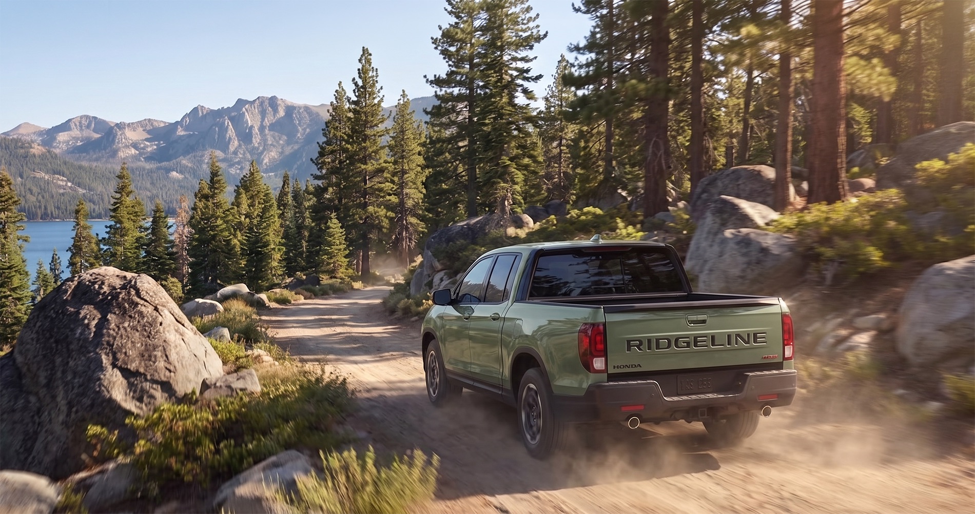 A green 2026 Honda Ridgeline driving through a dusty dirt road near Lake Tahoe with pine trees, granite boulders, and Sierra Nevada mountains in the background during warm afternoon light.