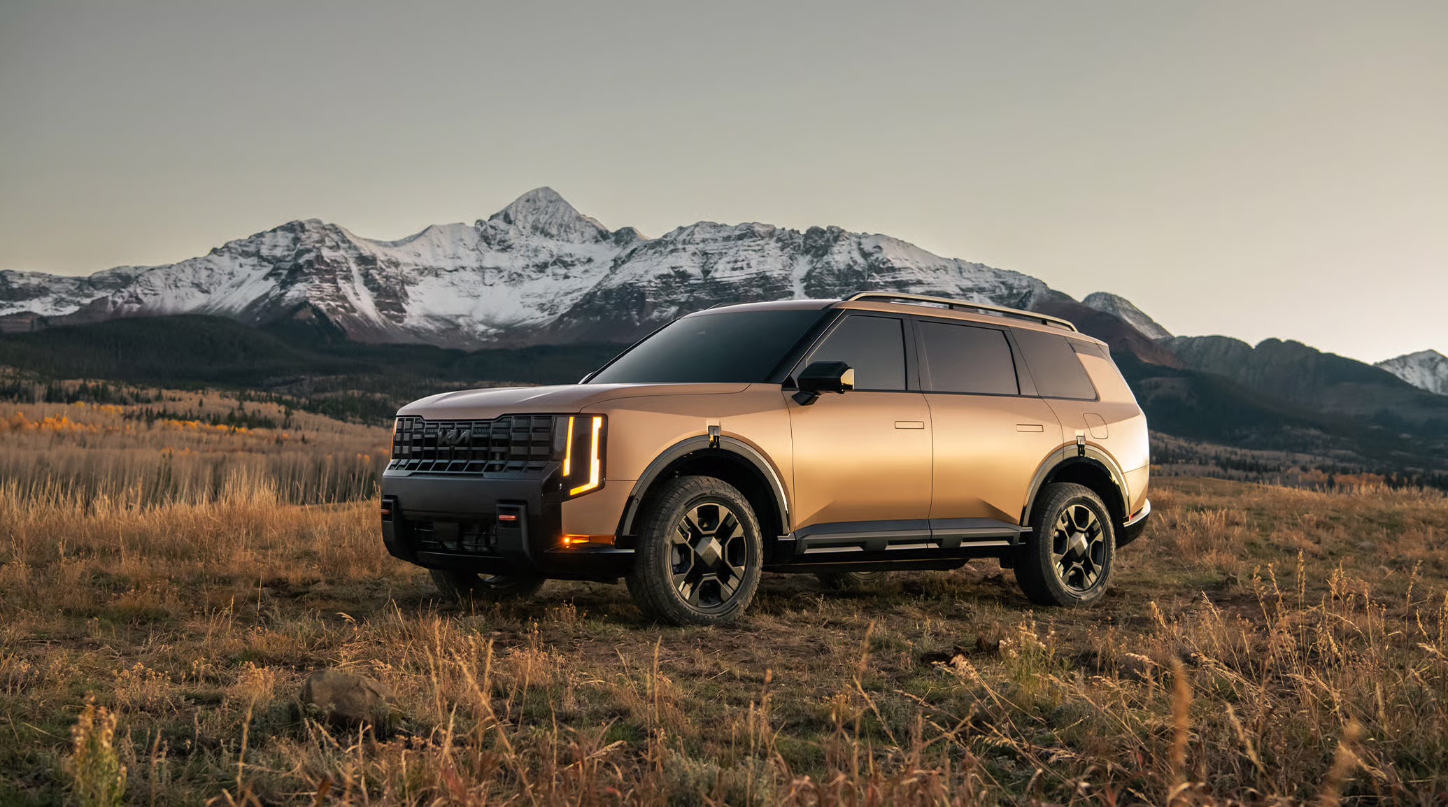 2026 Kia Telluride parked in an open mountain landscape at sunset with a bronze exterior, upright SUV styling, vertical LED lighting, rugged wheel design, and snow-covered peaks in the background.