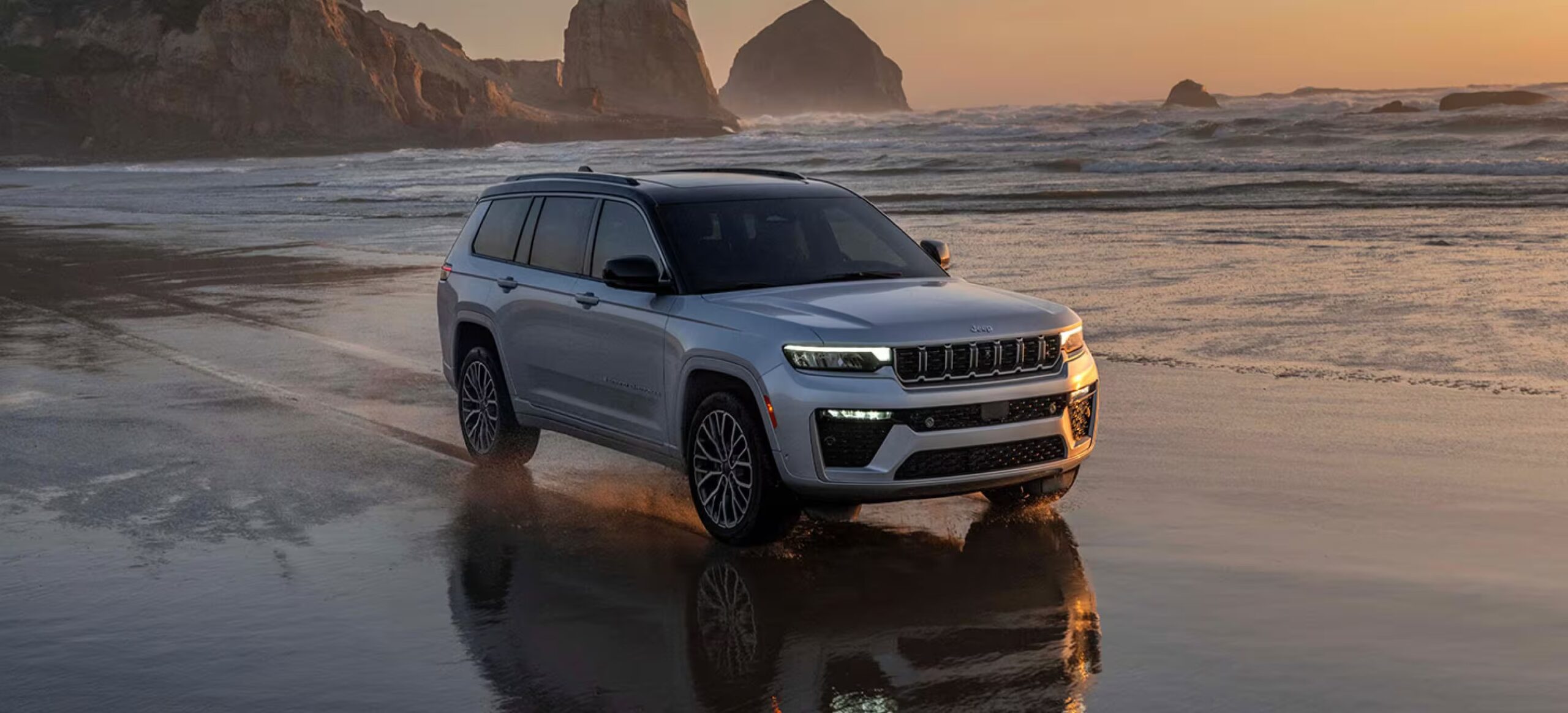 2026 Jeep Grand Cherokee driving along a wet beach at sunset with a white exterior, black roof accents, illuminated LED lighting, and updated seven-slot Jeep grille reflected against the shoreline and ocean backdrop.