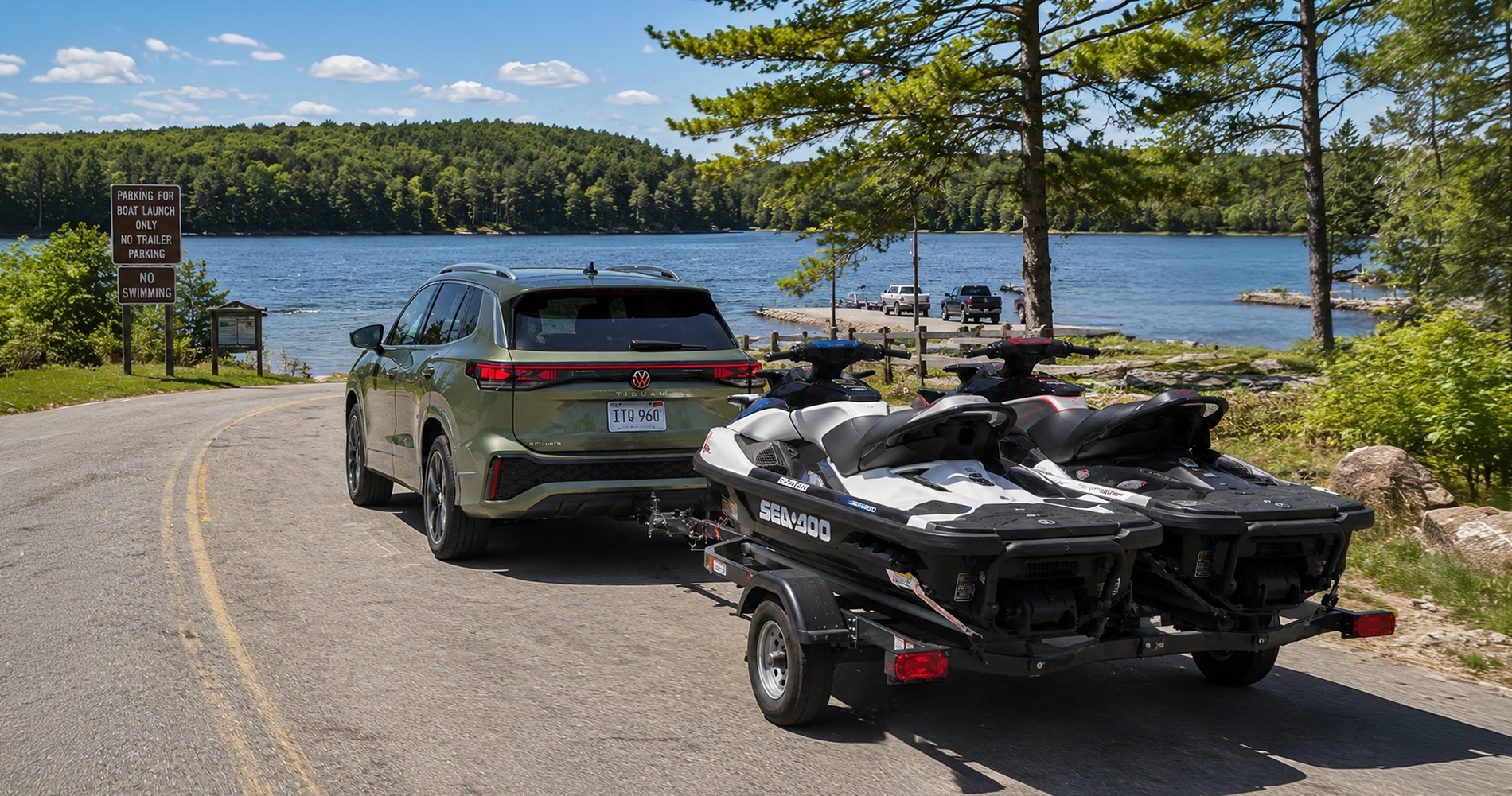 A green Volkswagen Tiguan towing two Sea-Doo jet skis toward a lakeside boat launch on a sunny summer day, surrounded by pine trees and calm water.