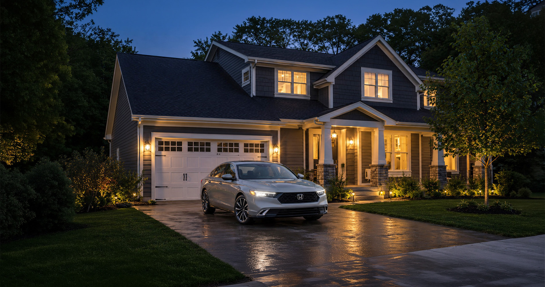 A silver 2026 Honda Accord parked in a residential driveway at dusk, with porch lights illuminating a gray two-story home and reflections on the wet pavement, illustrating the sedan’s refined styling and upscale presence in a suburban New York setting.