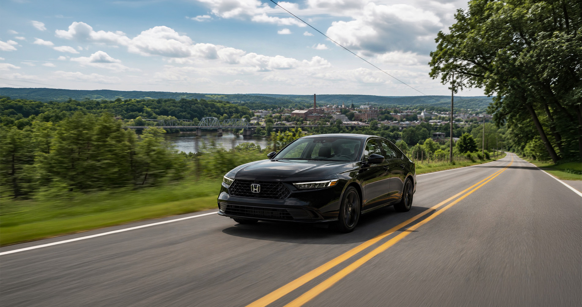 A black 2026 Honda Accord driving along a two-lane road overlooking the Hudson River and the city of Troy, New York, with a steel bridge and historic buildings visible in the background under a partly cloudy sky, highlighting the sedan’s sleek design and modern styling. html