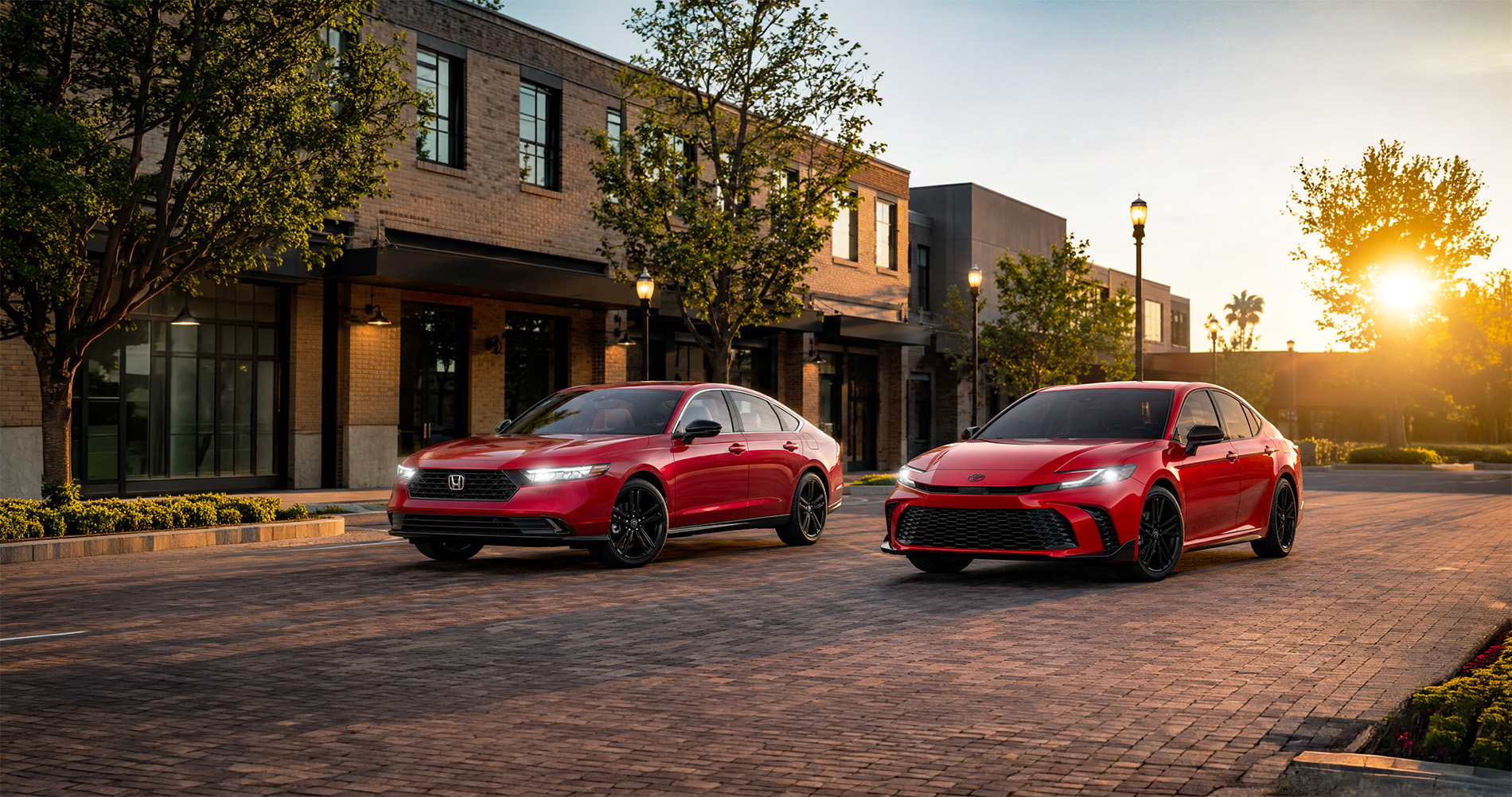 Two red sedans—a 2026 Honda Accord and a 2026 Toyota Camry—parked side by side on a brick-paved street in downtown Tracy, California, with modern buildings, trees, and lampposts illuminated by warm sunset light, highlighting the design differences between the Accord and Camry.