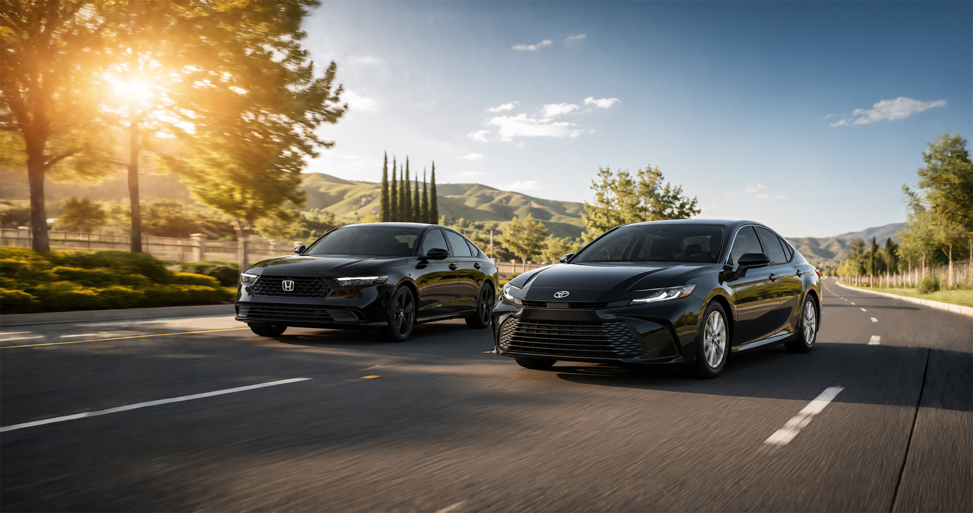 Two black sedans—a 2026 Honda Accord and a 2026 Toyota Camry—driving side by side on a tree-lined road surrounded by green hills under a clear blue sky, with sunlight filtering through the trees, showcasing the sleek styling and performance of both models.