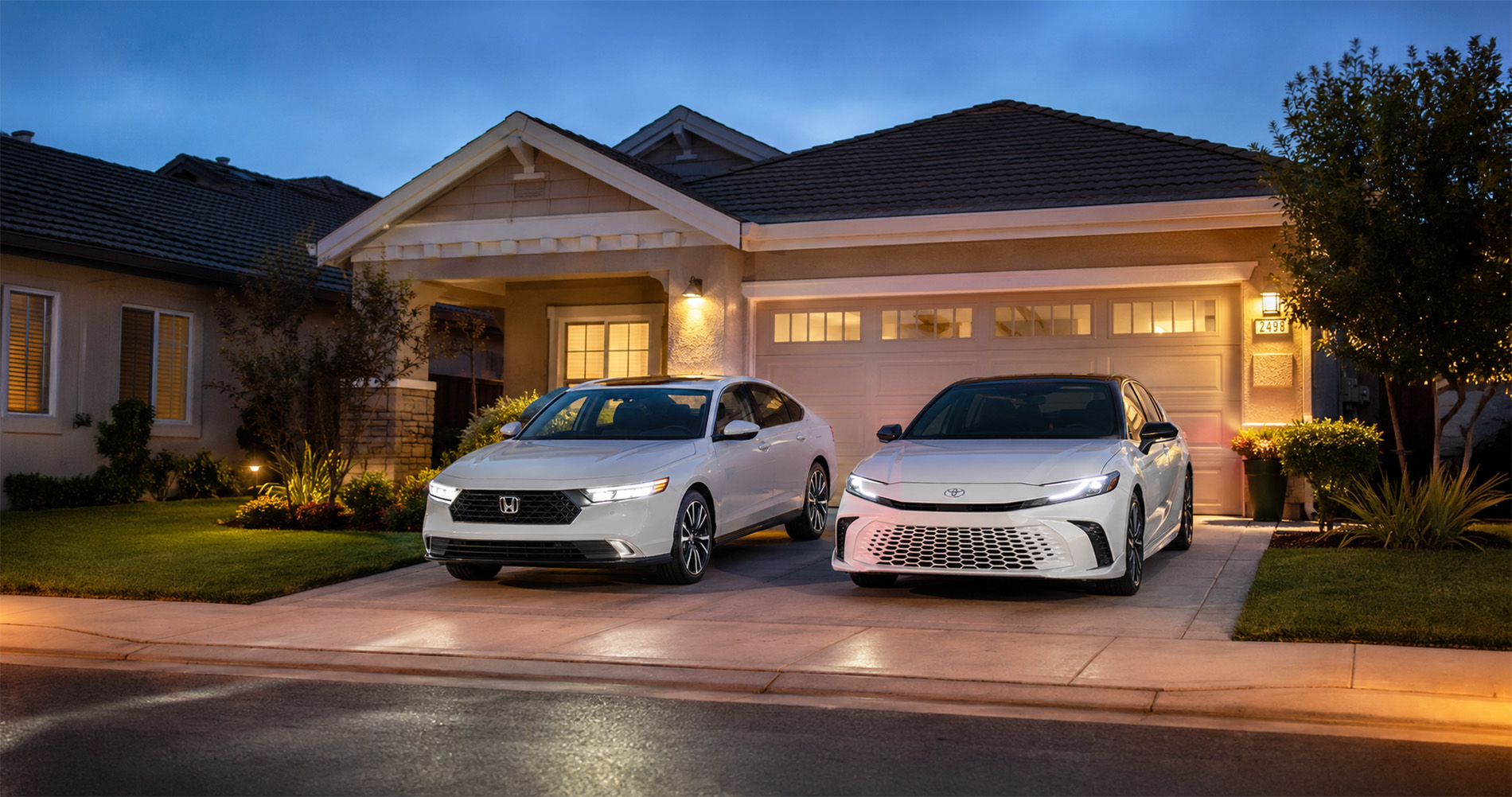 Two white sedans—a 2026 Honda Accord and a 2026 Toyota Camry—parked side by side in a residential driveway at dusk, with warm porch lights illuminating a modern home and reflections on the wet pavement, illustrating the refined design and everyday practicality of both vehicles.