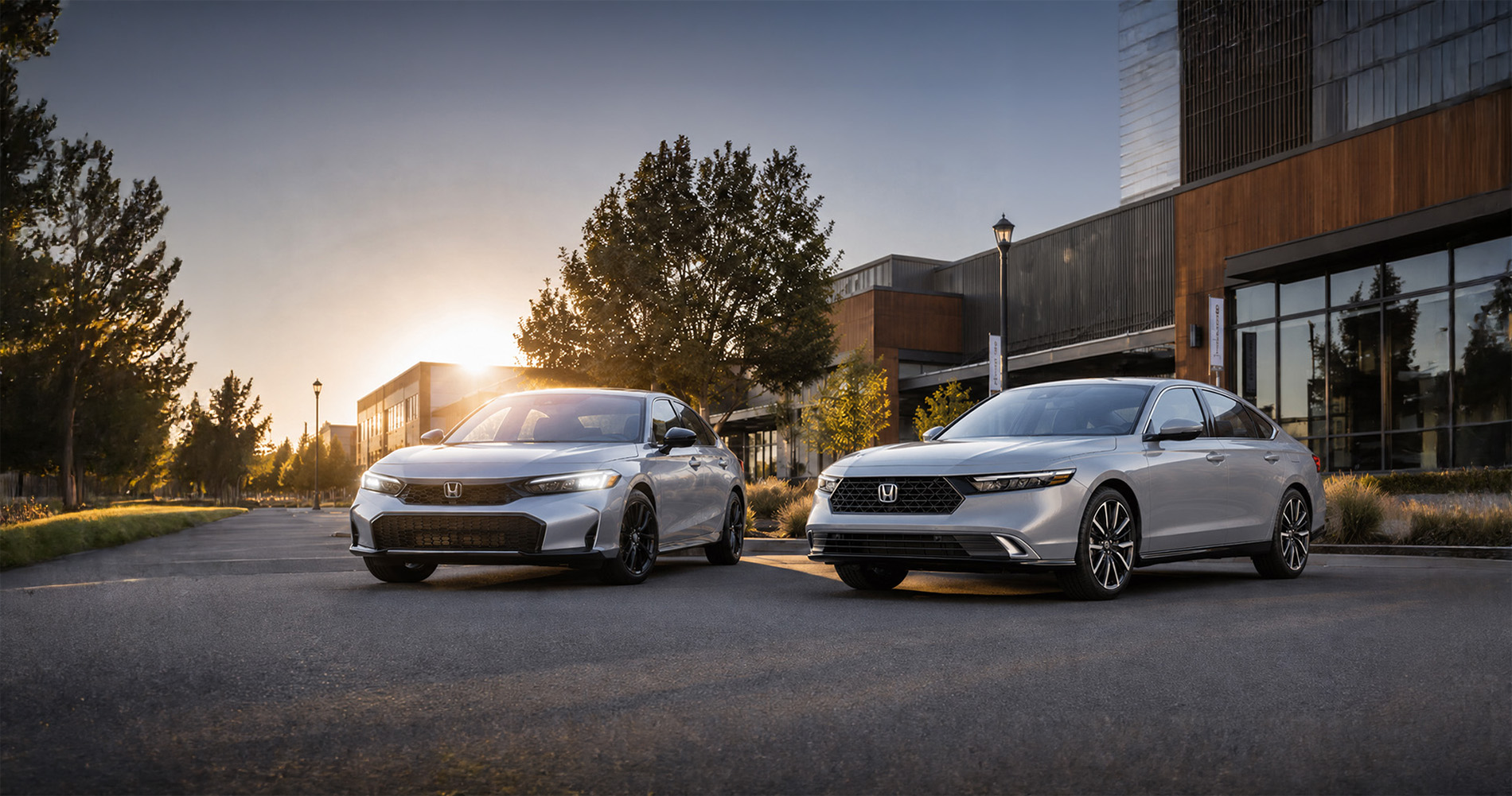 Two silver 2026 Honda sedans—a Civic and an Accord—parked side by side on a quiet street in front of modern buildings at sunset, with trees and lampposts casting long shadows, emphasizing the sleek styling and upscale design of both models.