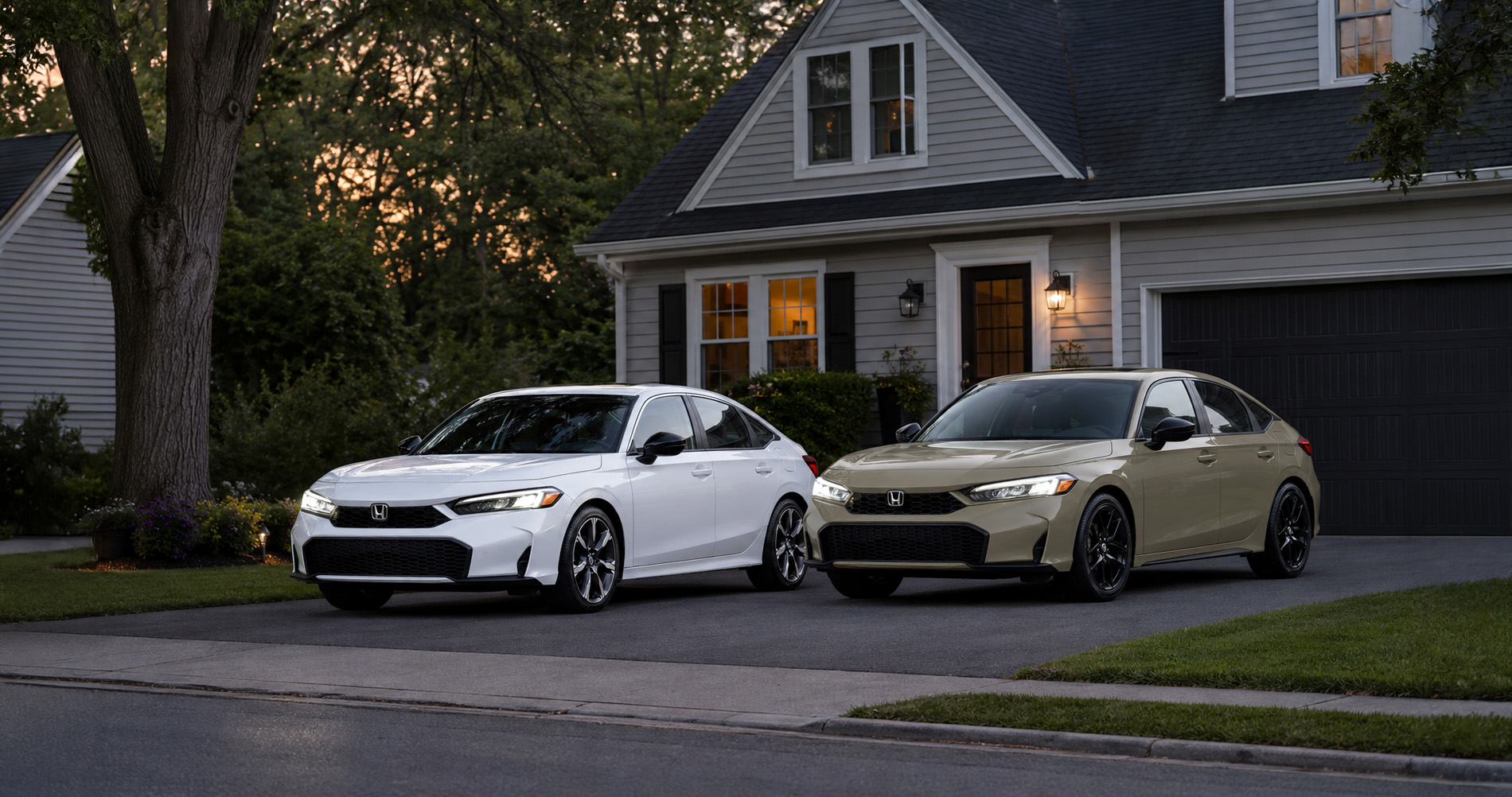 Two 2026 Honda Civic sedans in white and beige parked side by side in a residential driveway at dusk, with warm porch lights illuminating a gray two-story home, a large front-yard tree, and landscaped flower beds, showcasing the Civic’s refined styling in a suburban New York setting.