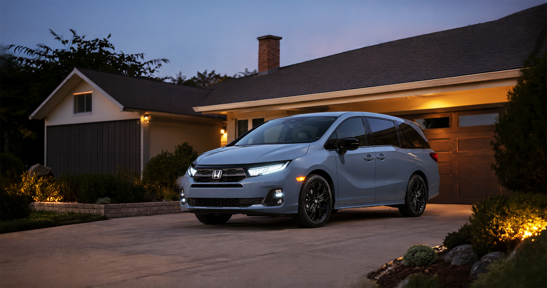 A light blue 2026 Honda Odyssey Sport-L parked in a residential driveway at dusk in El Cerrito, California, with headlights and porch lights glowing against a modern suburban home, highlighting the minivan’s sleek design and upscale exterior styling.