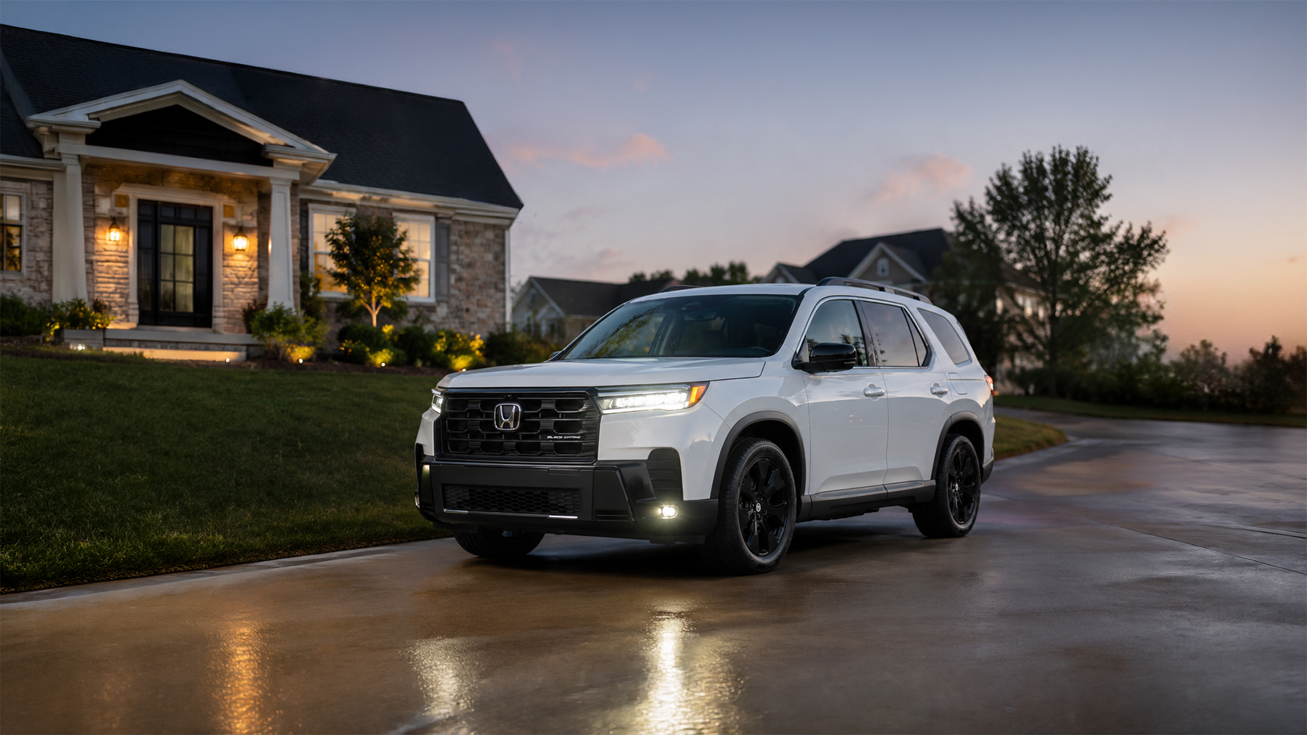 A white 2026 Honda Pilot Black Edition parked in a residential driveway at dusk, with porch lights illuminating a stone and siding home and trees in the background, showcasing the SUV’s refined styling and premium trim details