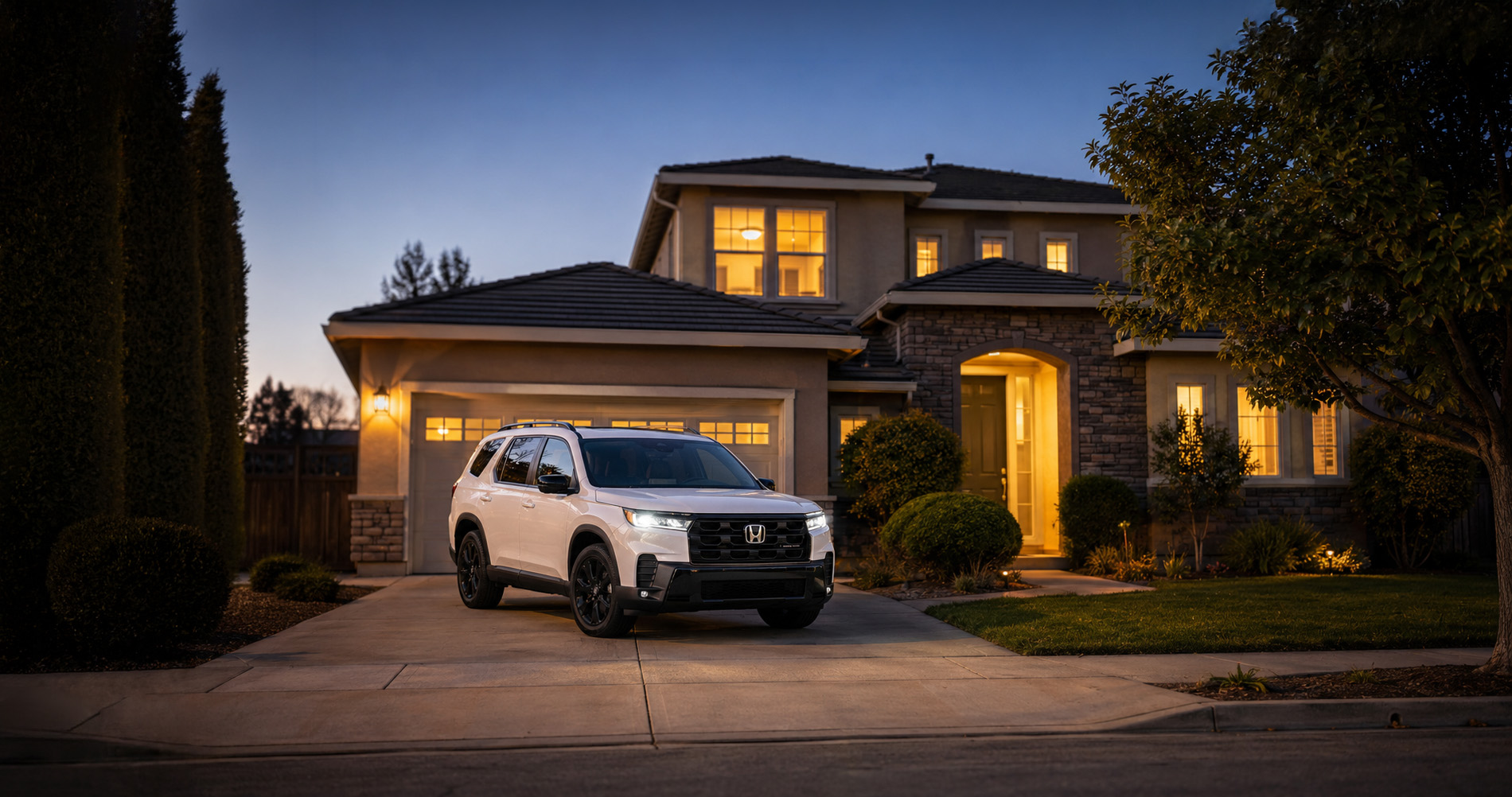 A white 2026 Honda Pilot Elite parked in the driveway of a modern two-story home at dusk in Pittsburg, California, with warm house lights illuminating the exterior and trees framing the scene, showcasing the SUV’s upscale styling and family-friendly appeal.
