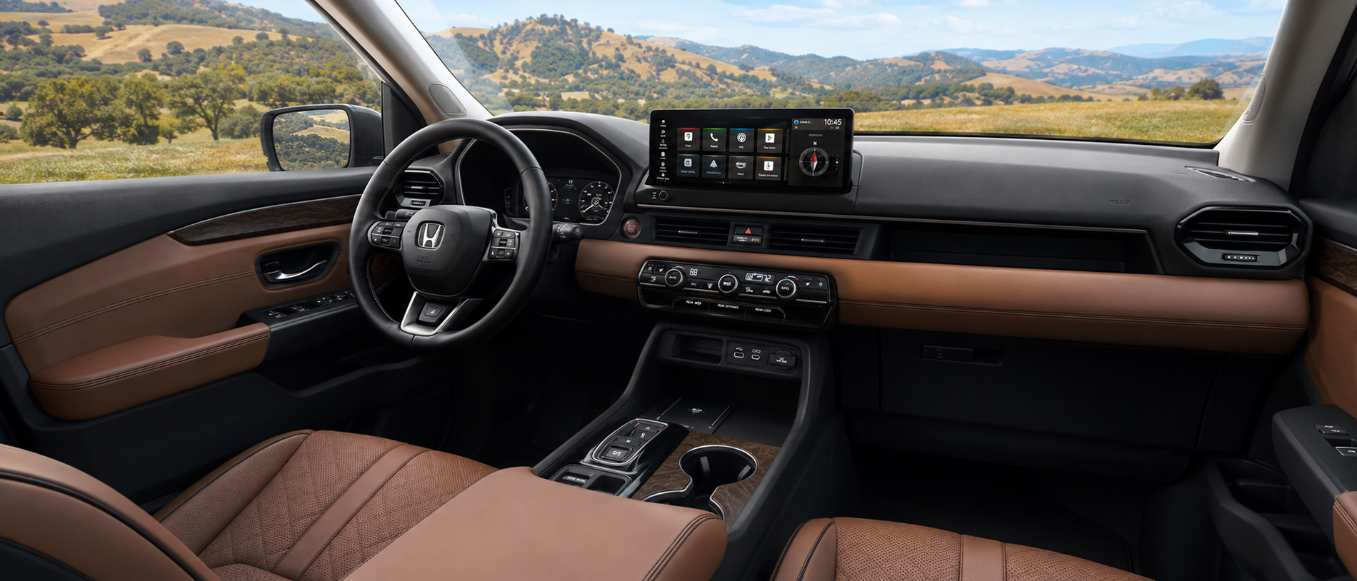 The interior of a 2026 Honda Pilot featuring brown leather seats, wood trim, and a large touchscreen display showing navigation and apps, with a scenic view of grassy hills and blue skies visible through the windshield, emphasizing the SUV’s premium craftsmanship and technology.