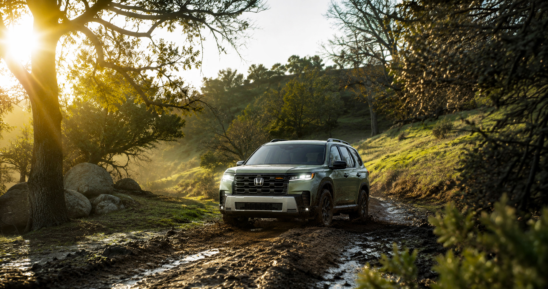 A green 2026 Honda Pilot TrailSport driving through a muddy forest trail surrounded by trees and sunlight filtering through branches near Pittsburg, California, highlighting the SUV’s rugged design and off-road performance.