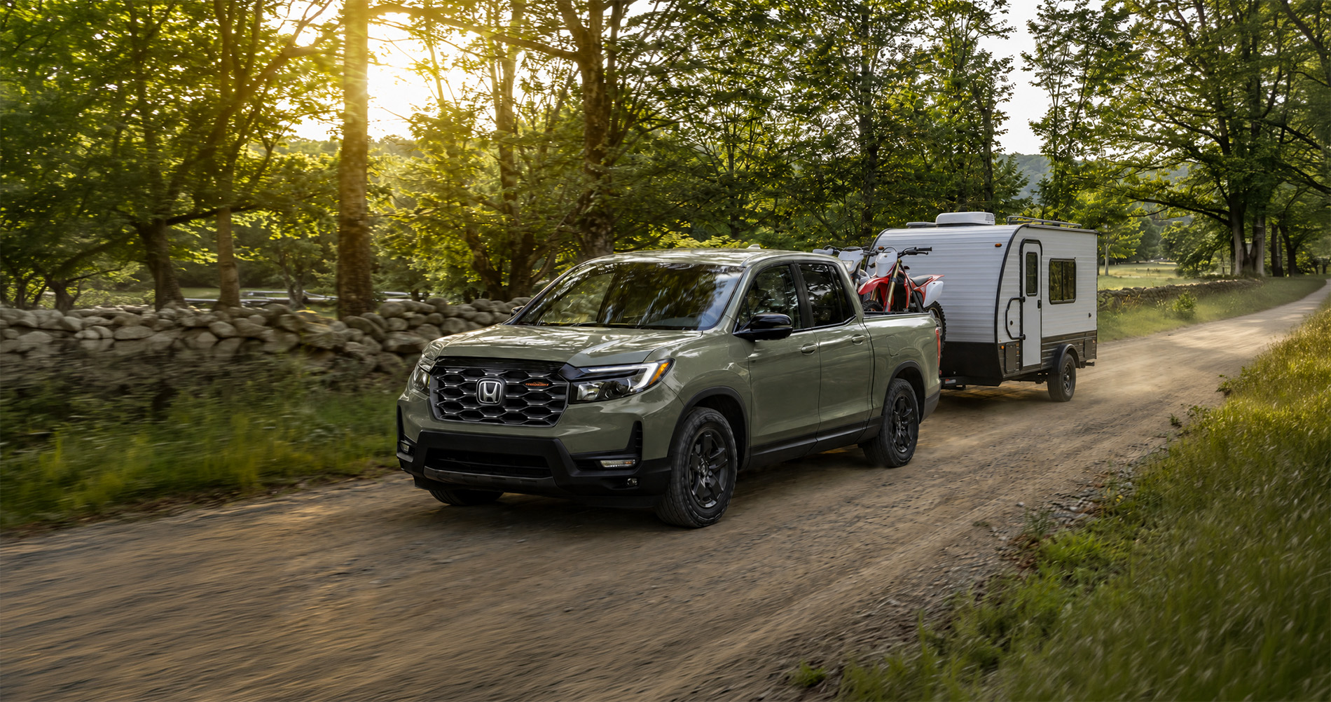2026 Honda Ridgeline TrailSport towing a small camper with dirt bikes loaded in the bed in a rural setting.