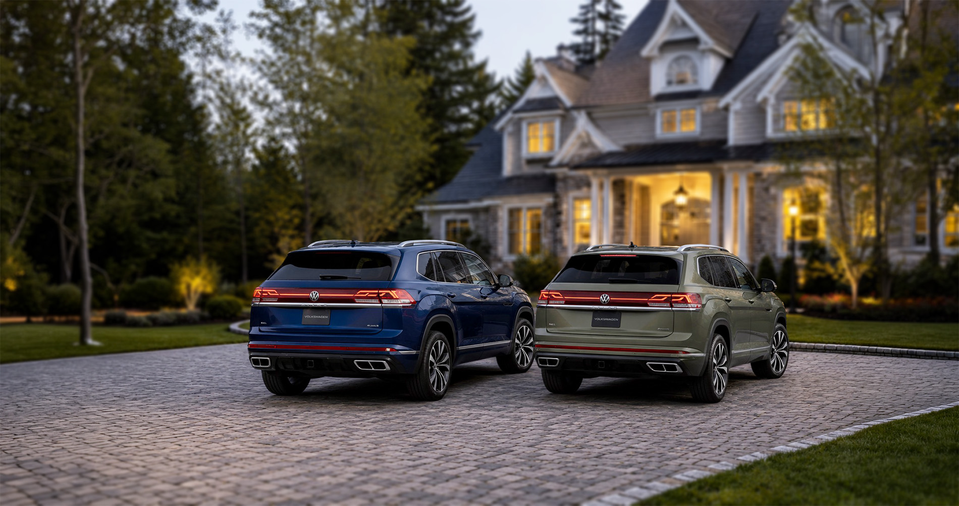 Two Volkswagen SUVs—a blue Atlas and a green Atlas Cross Sport—parked side by side on a cobblestone driveway in front of a large, well-lit home surrounded by trees in Easton, Massachusetts.