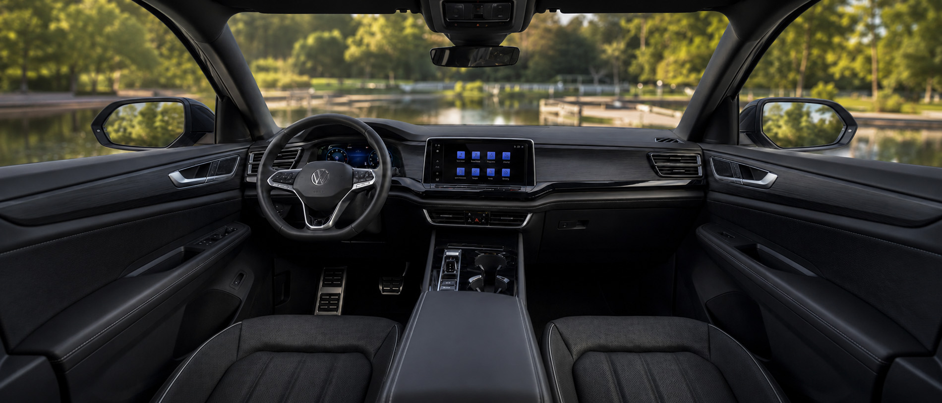 The interior of a 2026 Volkswagen Atlas featuring black leather seats, a digital instrument cluster, and a large touchscreen display, with a scenic view of a red barn and stone bridge over a calm river visible through the windshield.