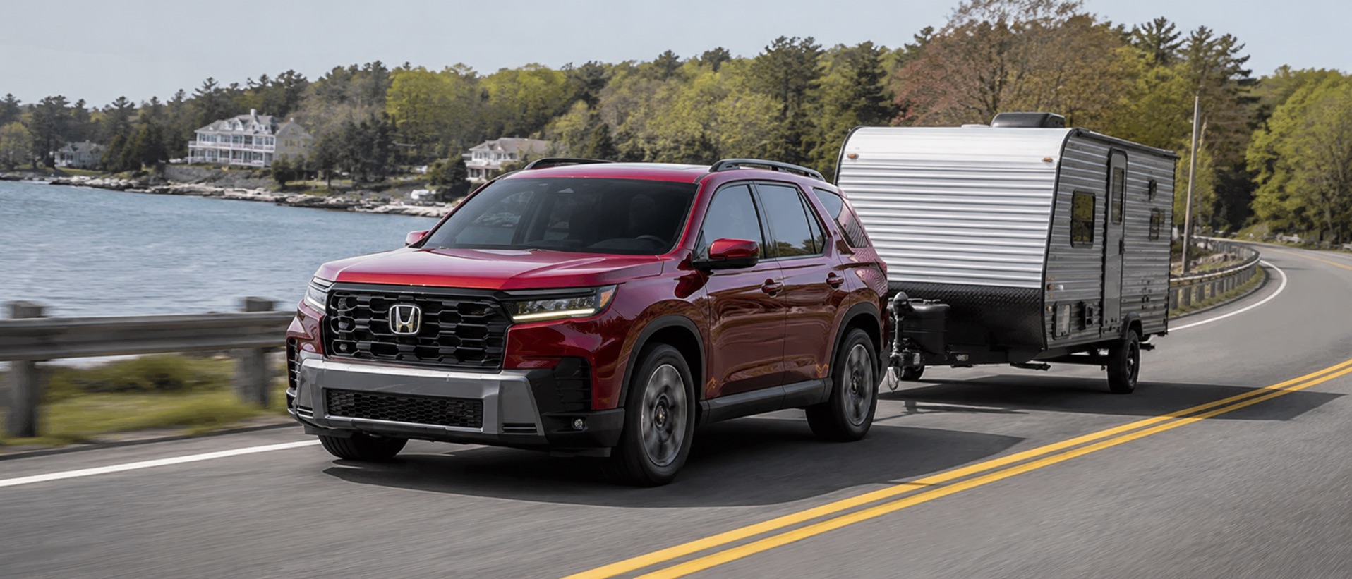 Red 2026 Honda Pilot towing a small camper trailer along a scenic southeastern Massachusetts waterfront road