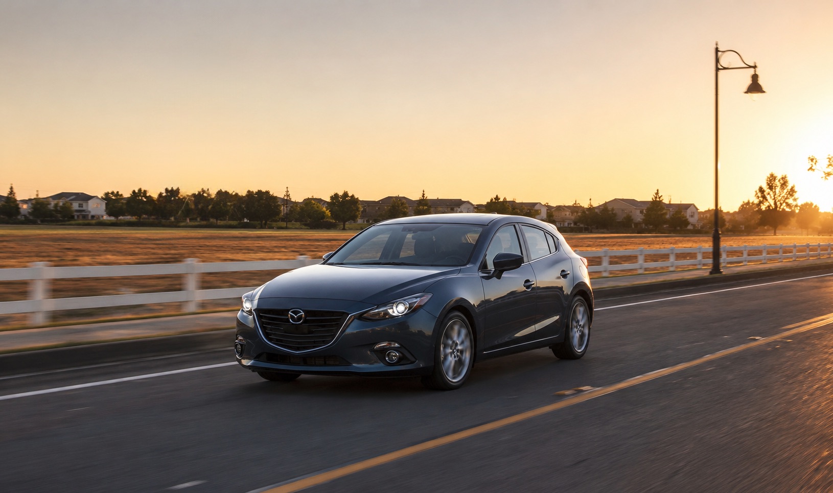 Dark blue Mazda3 hatchback driving along a road near Tracy, California at sunset, with open farmland, a white ranch fence, suburban homes, and the Tracy water tower visible in the background under warm golden-hour light.