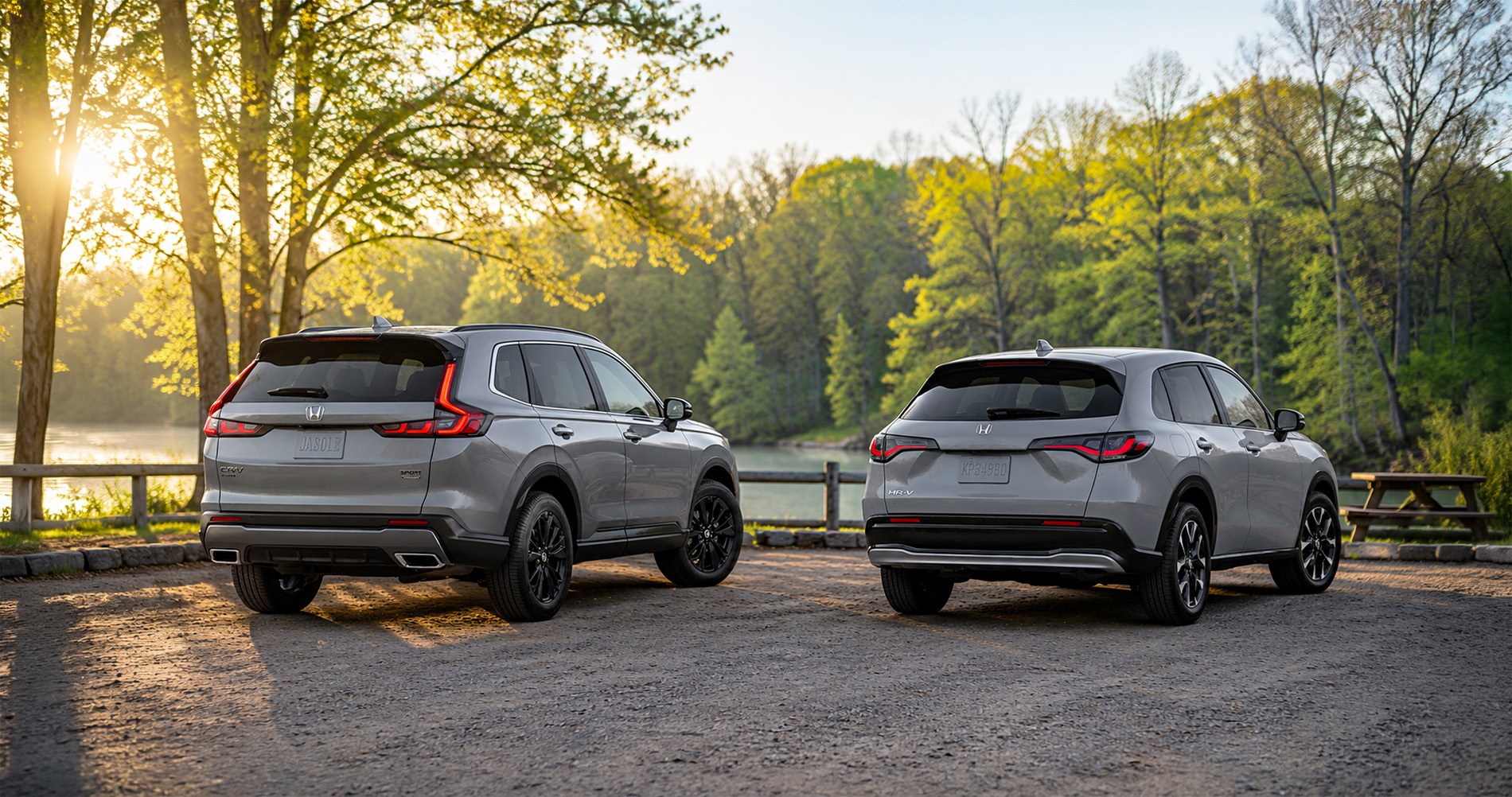 Silver Honda CR-V and Honda HR-V SUVs parked lakeside at sunset, showing rear styling and taillight design.