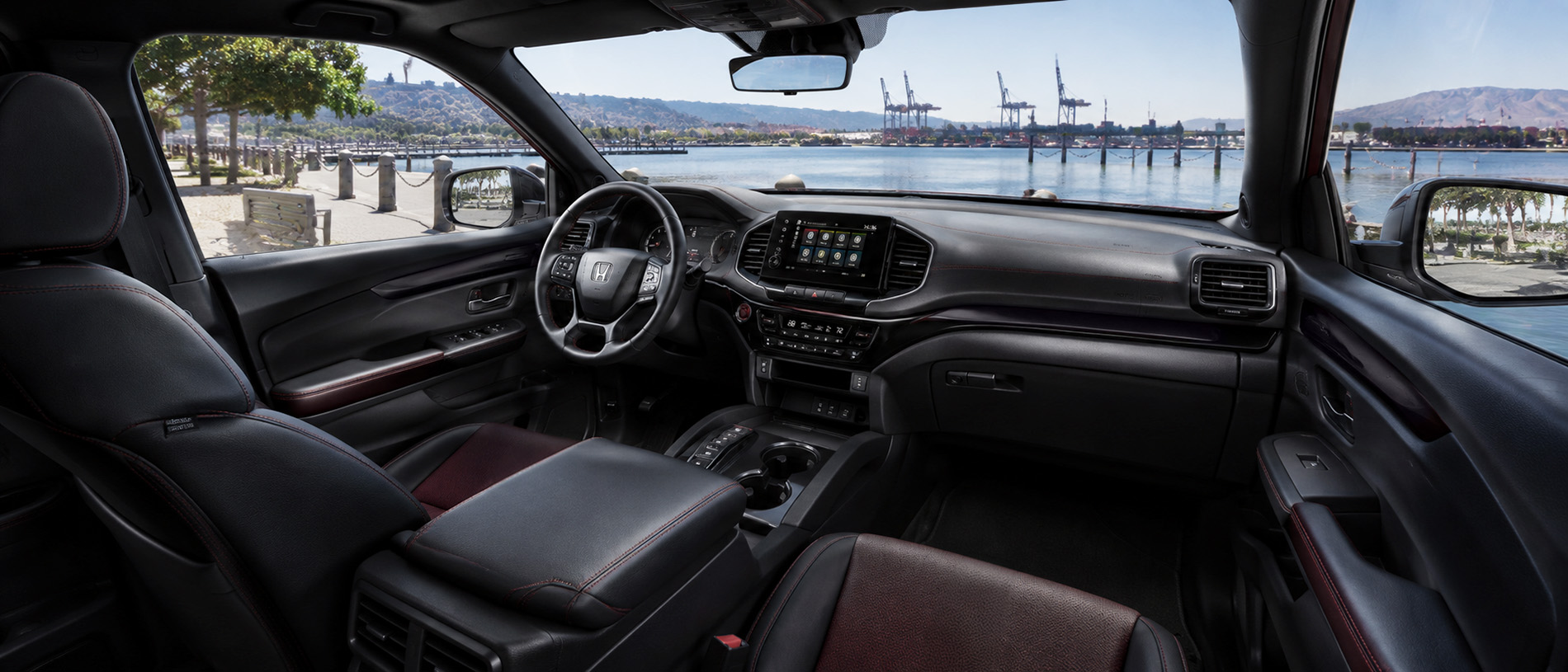 Black leather cabin with red stitching and touchscreen display, overlooking waterfront cranes and hills.