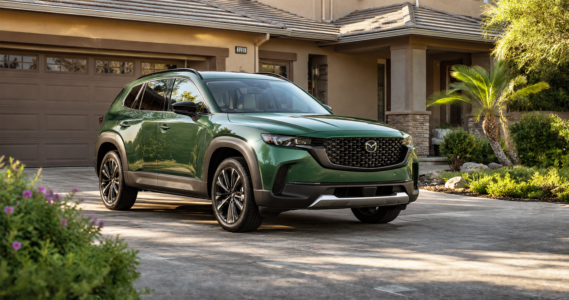 Green Mazda CX-50 SUV parked in driveway at sunrise highlighting sleek design and black grille accents.