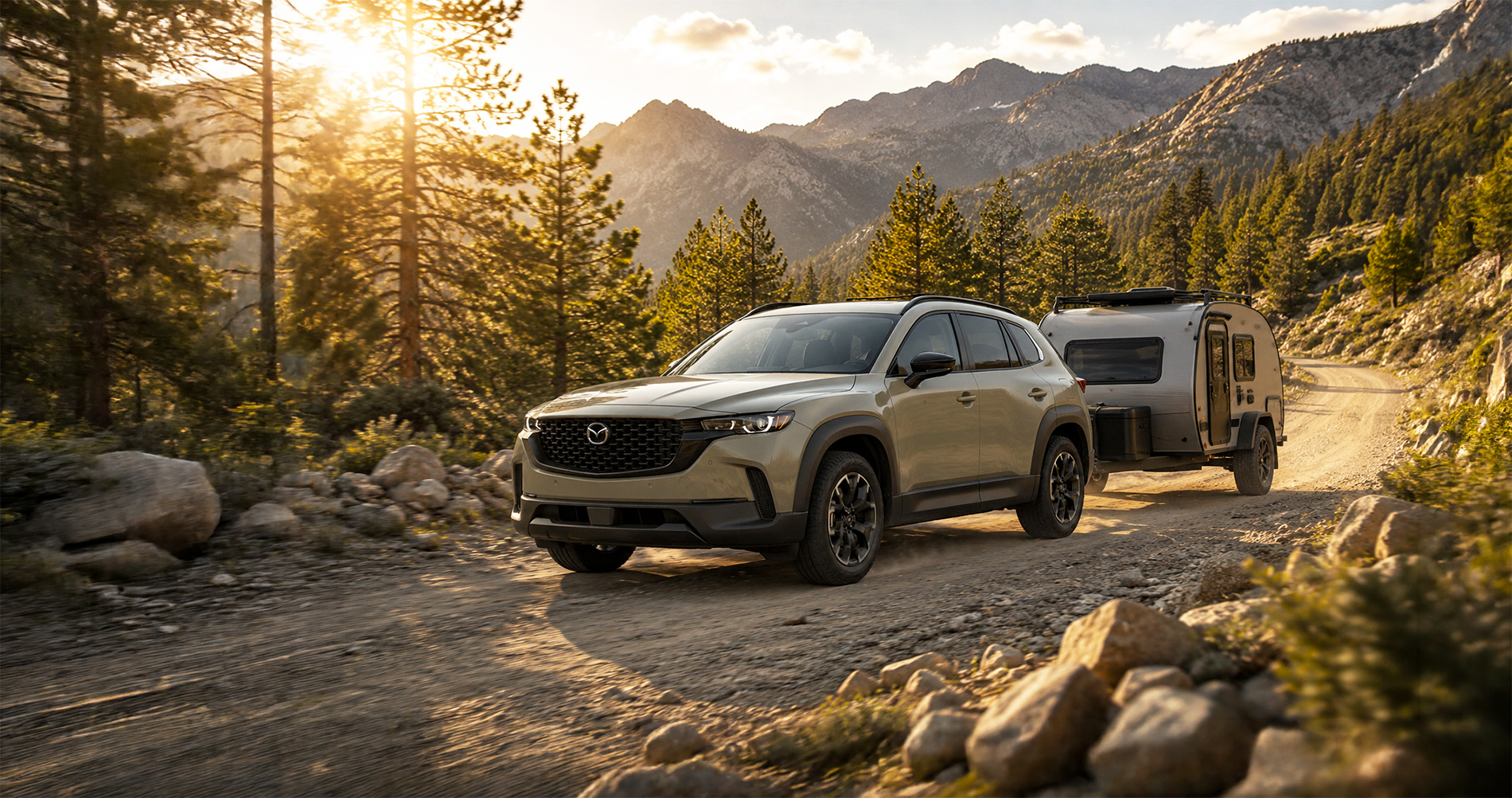 Tan Mazda CX-50 towing small camper on dirt mountain road at sunset emphasizing off road capability.