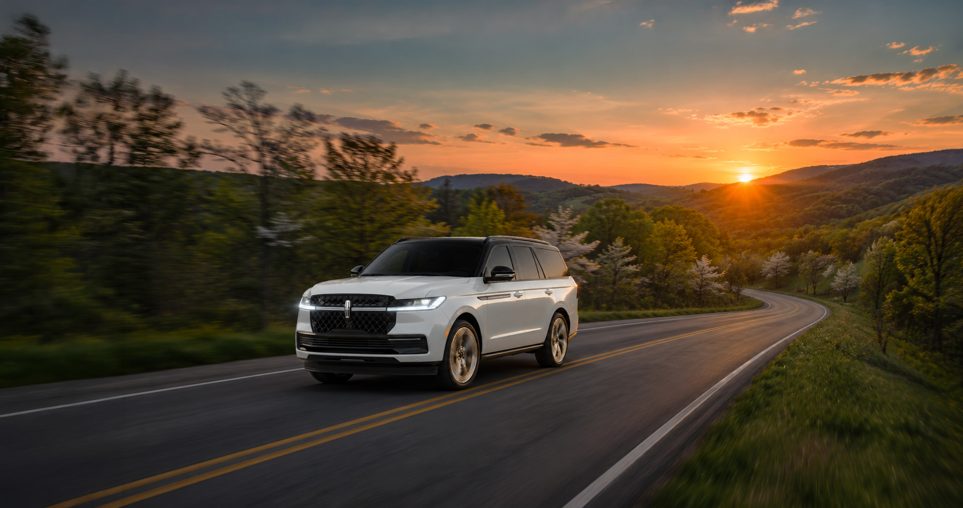 White Lincoln Navigator driving on a winding forest road at sunset.