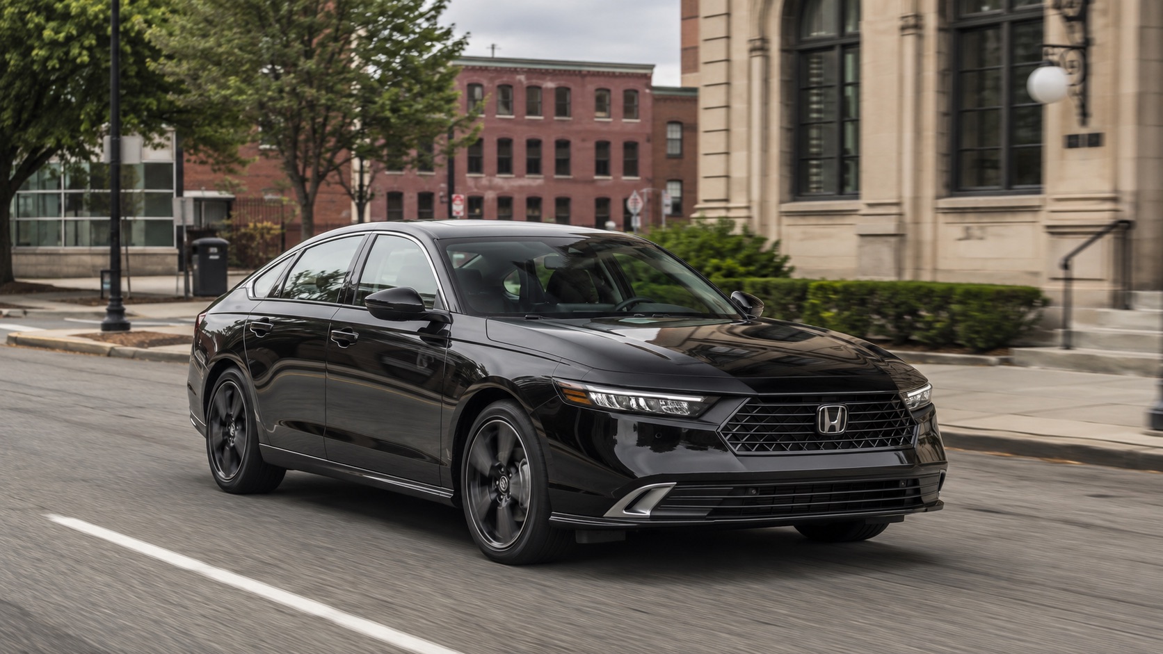 Black 2026 Honda Accord Sport Touring Hybrid driving through Brockton, Massachusetts with historic brick and civic-style architecture in the background.