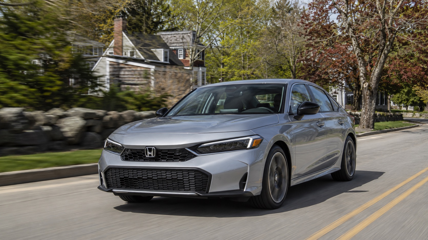 Silver 2026 Honda Civic Touring Hybrid driving through a tree-lined Raynham, Massachusetts neighborhood with classic New England scenery.