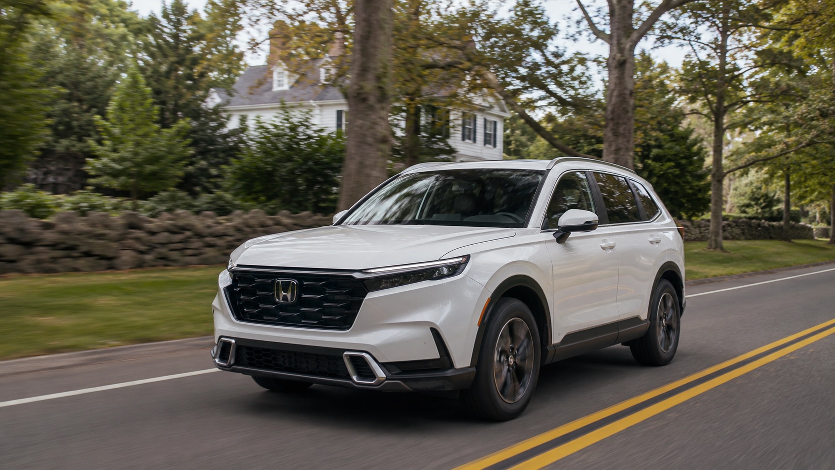 White 2026 Honda CR-V Touring Hybrid driving through Easton, Massachusetts on a wooded road lined with stone walls and New England homes.
