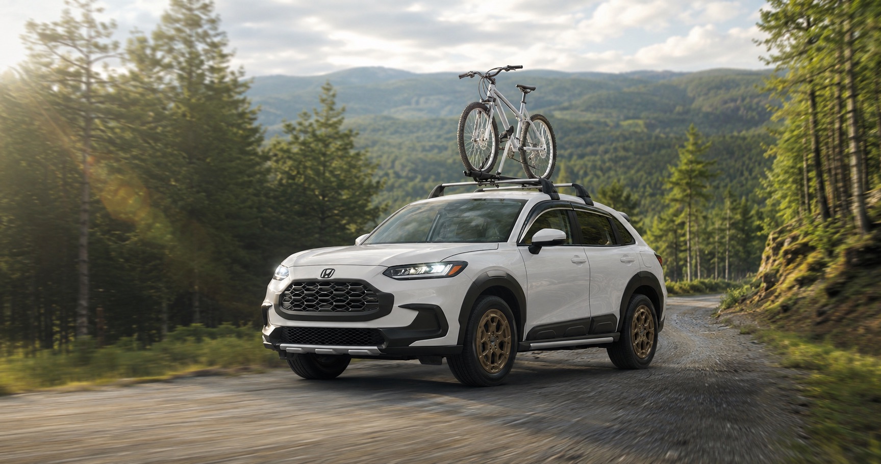 White Honda HR‑V with bicycle mounted on roof driving on forest road at sunrise emphasizing outdoor capability and rugged styling.