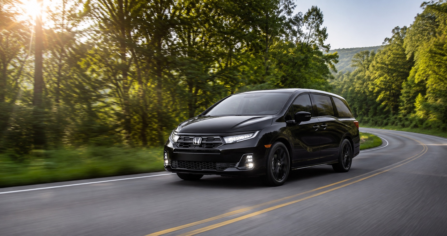 Black Honda Odyssey driving on winding forest road at sunrise emphasizing sleek design and smooth performance.